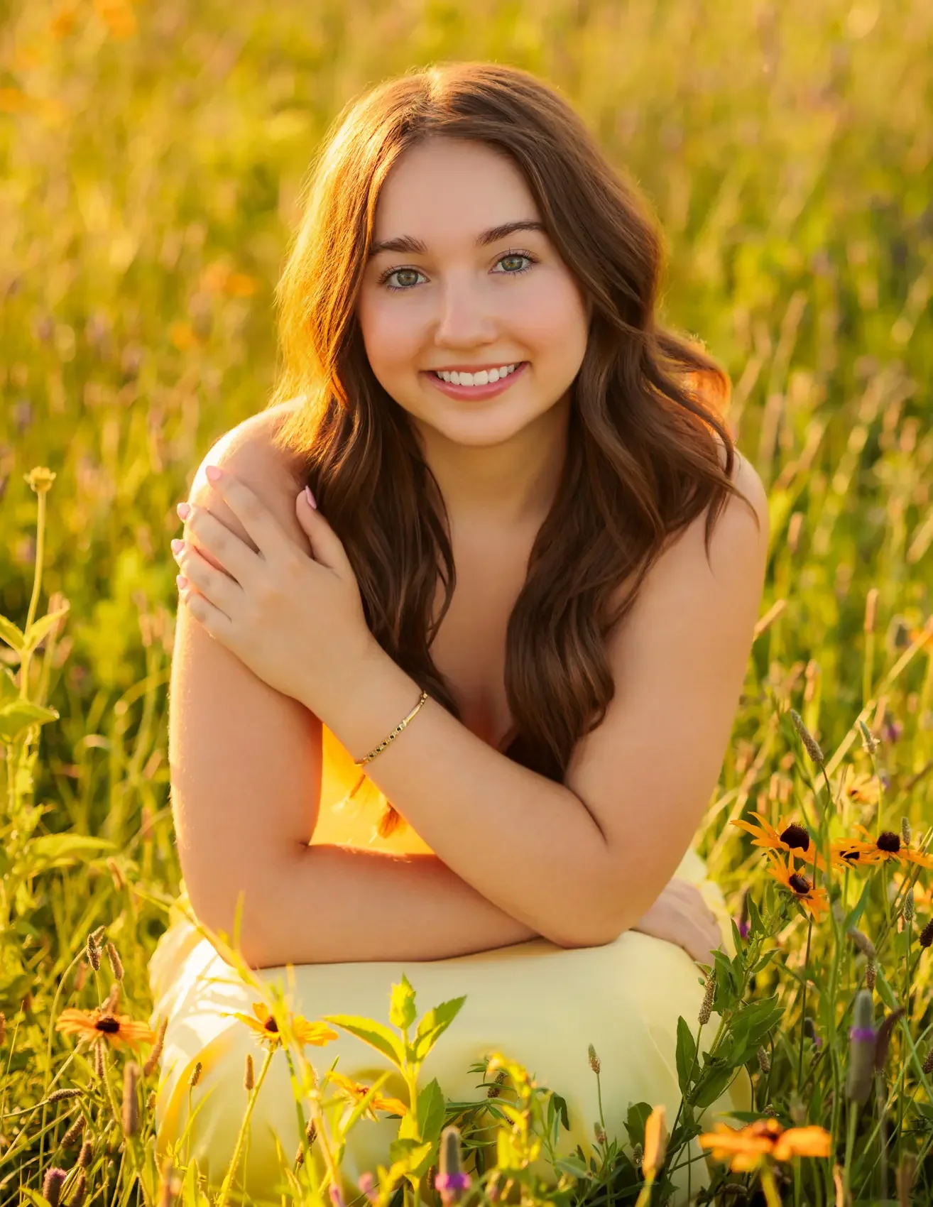 Senior girl with long brown hair and blue eyes smiling in a field of yellow flowers during sunset, wearing a yellow dress with arms crossed over her chest, posing for senior pictures in Des Moines.