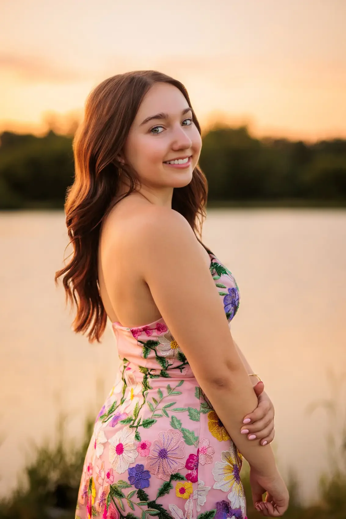 High school senior girl with long brown hair in a colorful floral dress, standing outdoors at sunset.