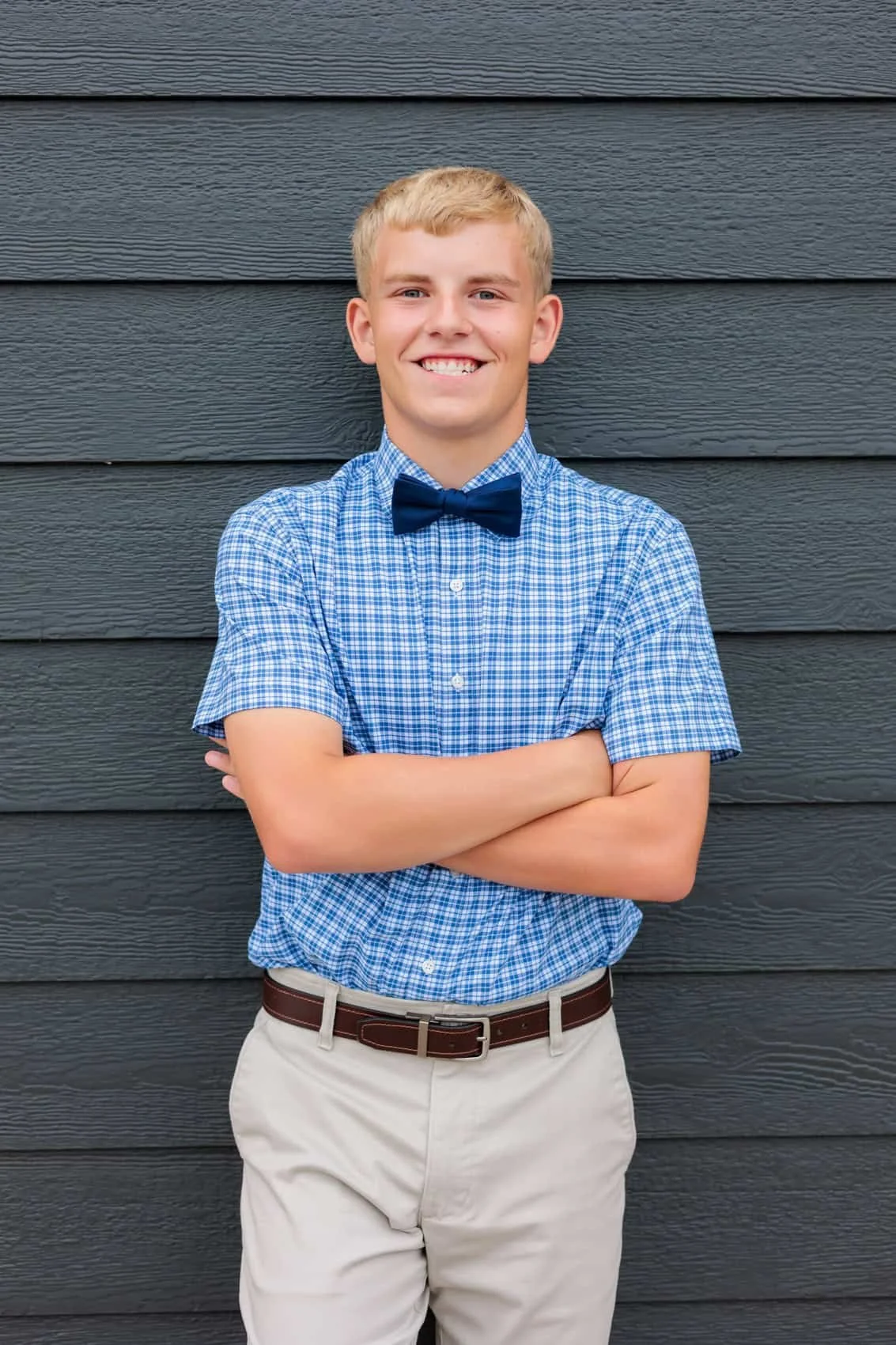  High school senior boy with blond hair smiling, wearing a blue checkered shirt, dark blue bow tie, beige pants, and a brown belt, standing with arms crossed in front of a dark gray wooden wall.