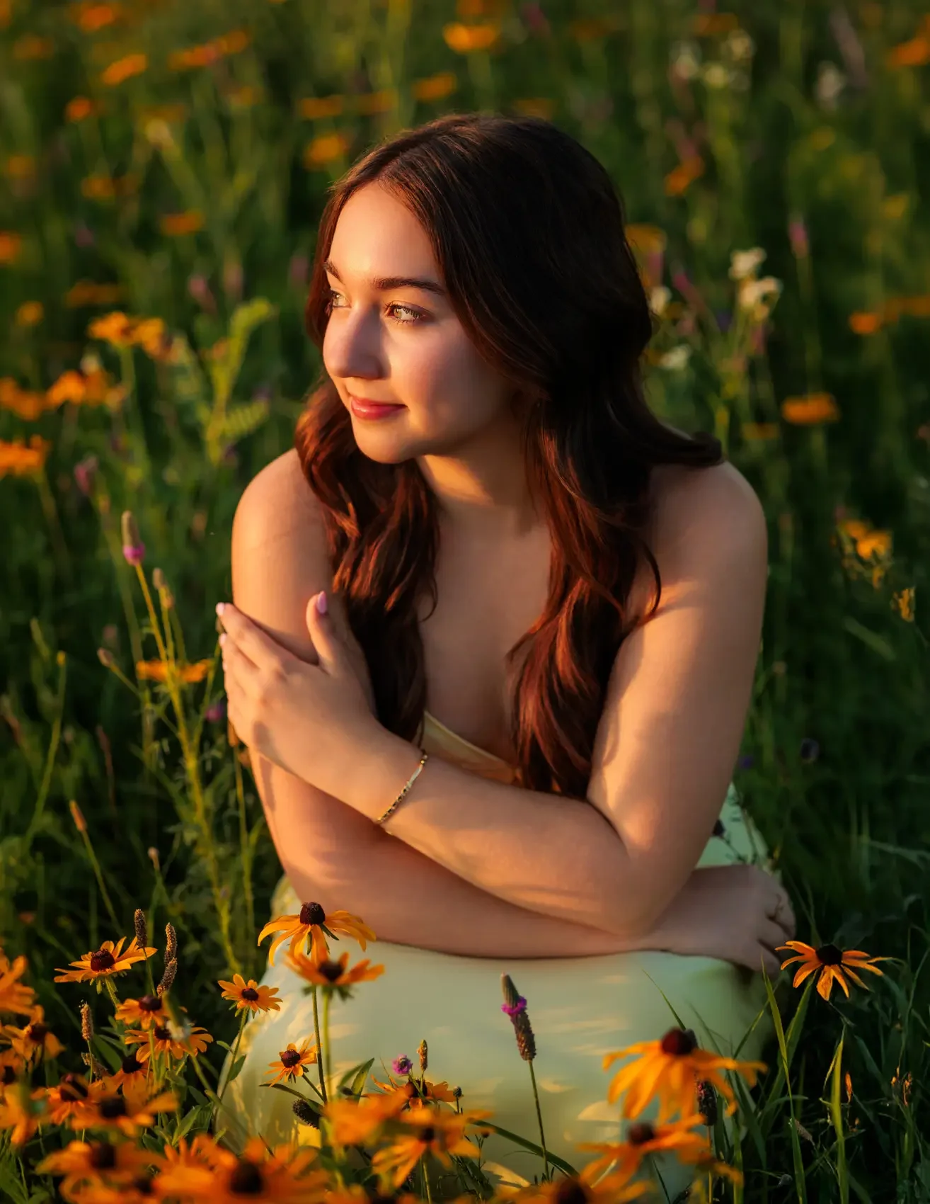 Senior girl with brown hair in loose waves sitting among orange flowers in a field, illuminated by warm, golden sunlight, looking thoughtfully to the side with arms crossed.