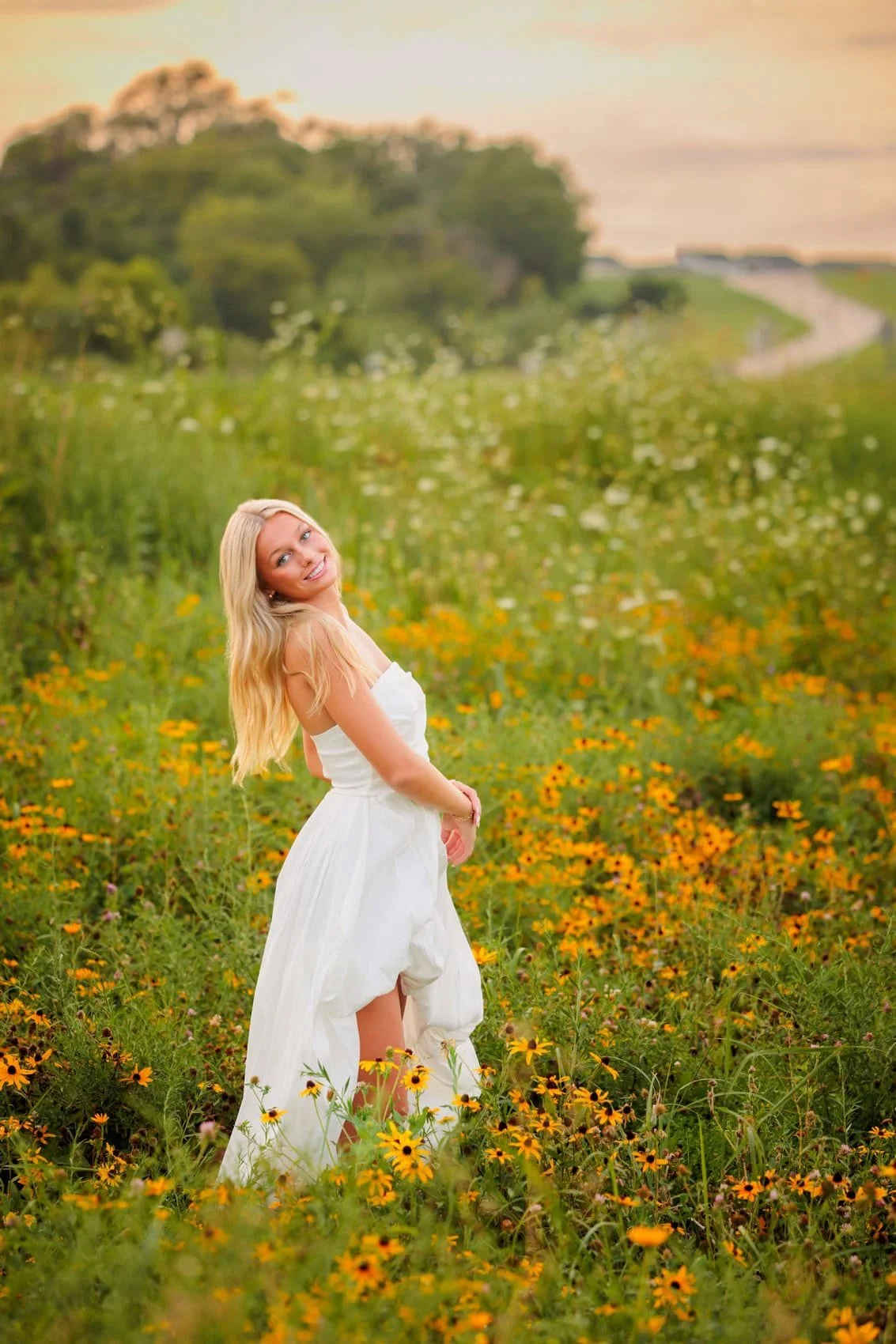Senior girl with long blonde hair wearing a white dress standing in a field of yellow flowers.