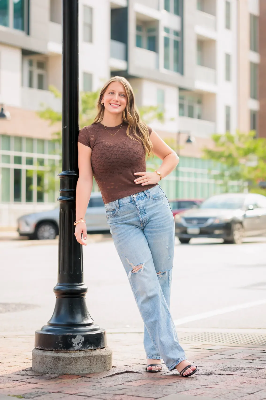 Senior girl with long brown hair, wearing a brown lace shirt, ripped jeans, and black sandals, leaning against a black street lamp on a city sidewalk in the Court Avenue distict of Des Moines during her senior photo session.