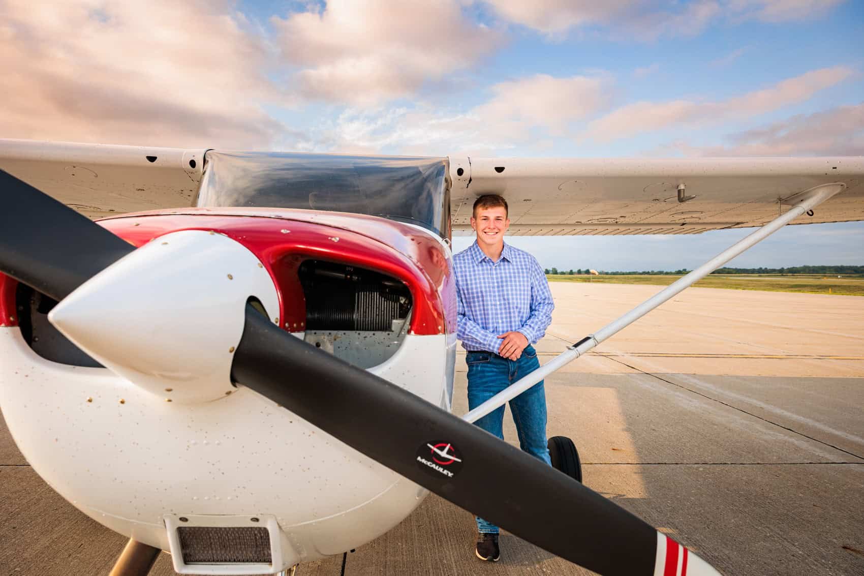  High school senior guy in a blue checkered shirt and jeans standing next to a small airplane on an airport tarmac, with the airplane's propeller prominently in the foreground.