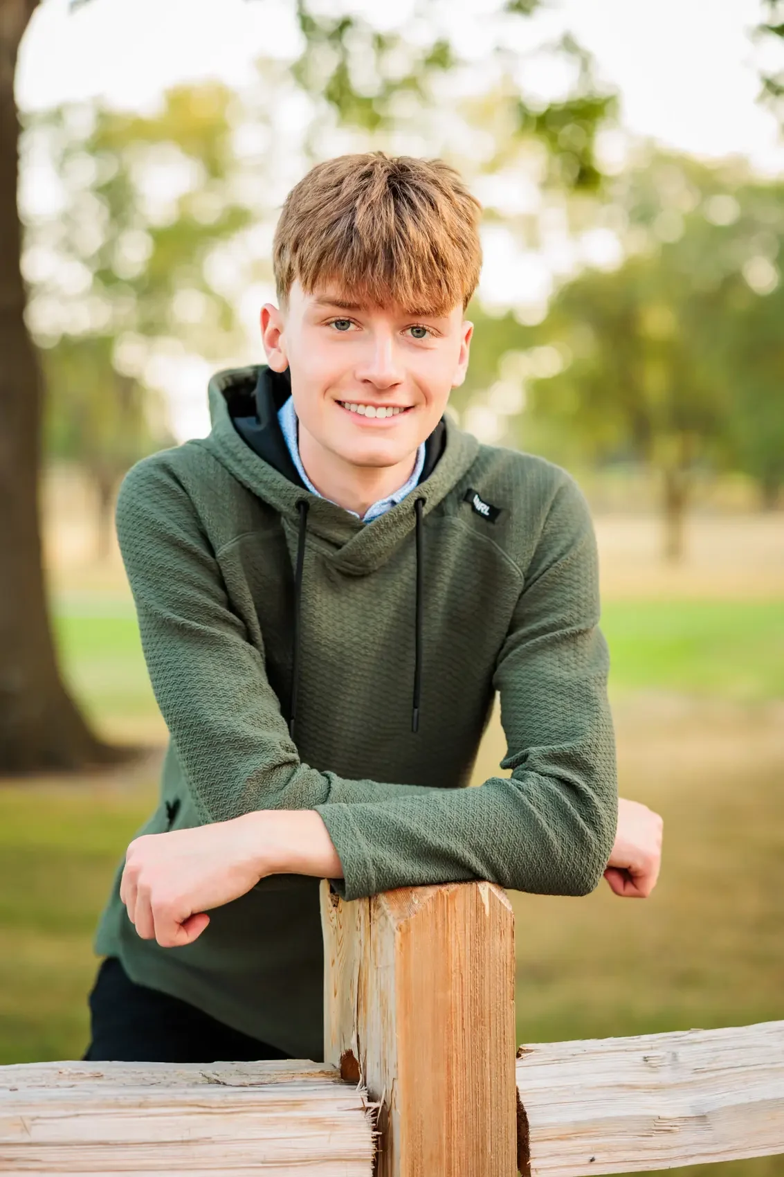 Senior boy with reddish hair and light skin wearing a green hoodie, smiling and leaning on a wooden fence in a park with trees in the background.