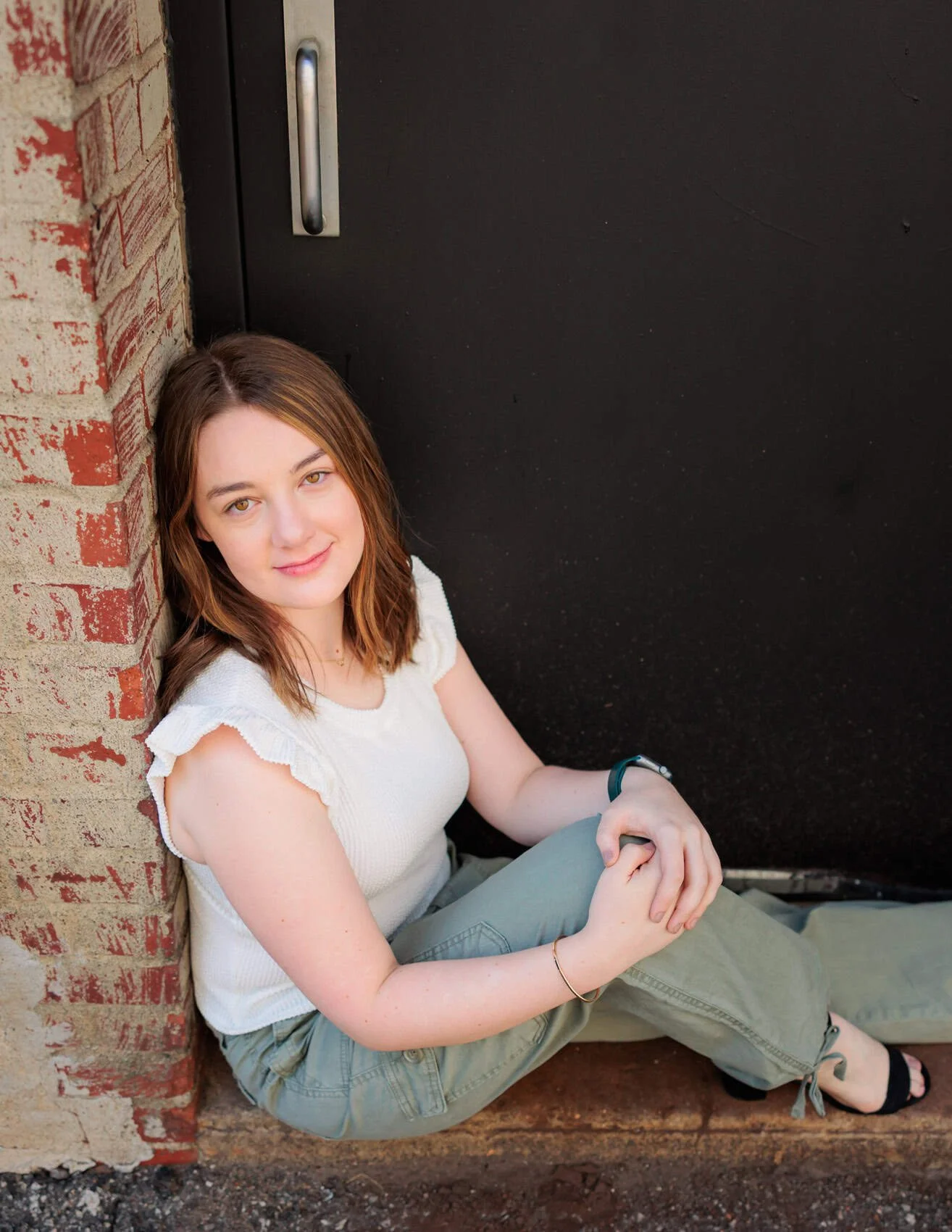 High school senior girl with shoulder-length brown hair and light skin, sitting on the ground with her back against a brick wall and a black door behind her. 