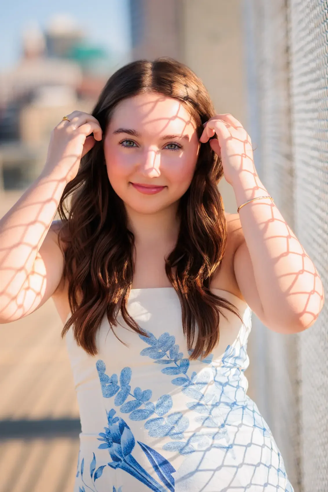 A young woman with long, wavy brown hair and blue eyes on a sunny day, standing outdoors near a wall with a wire fence casting shadows on her face and arms. She is wearing a white dress with a blue floral pattern and smiling at the camera.