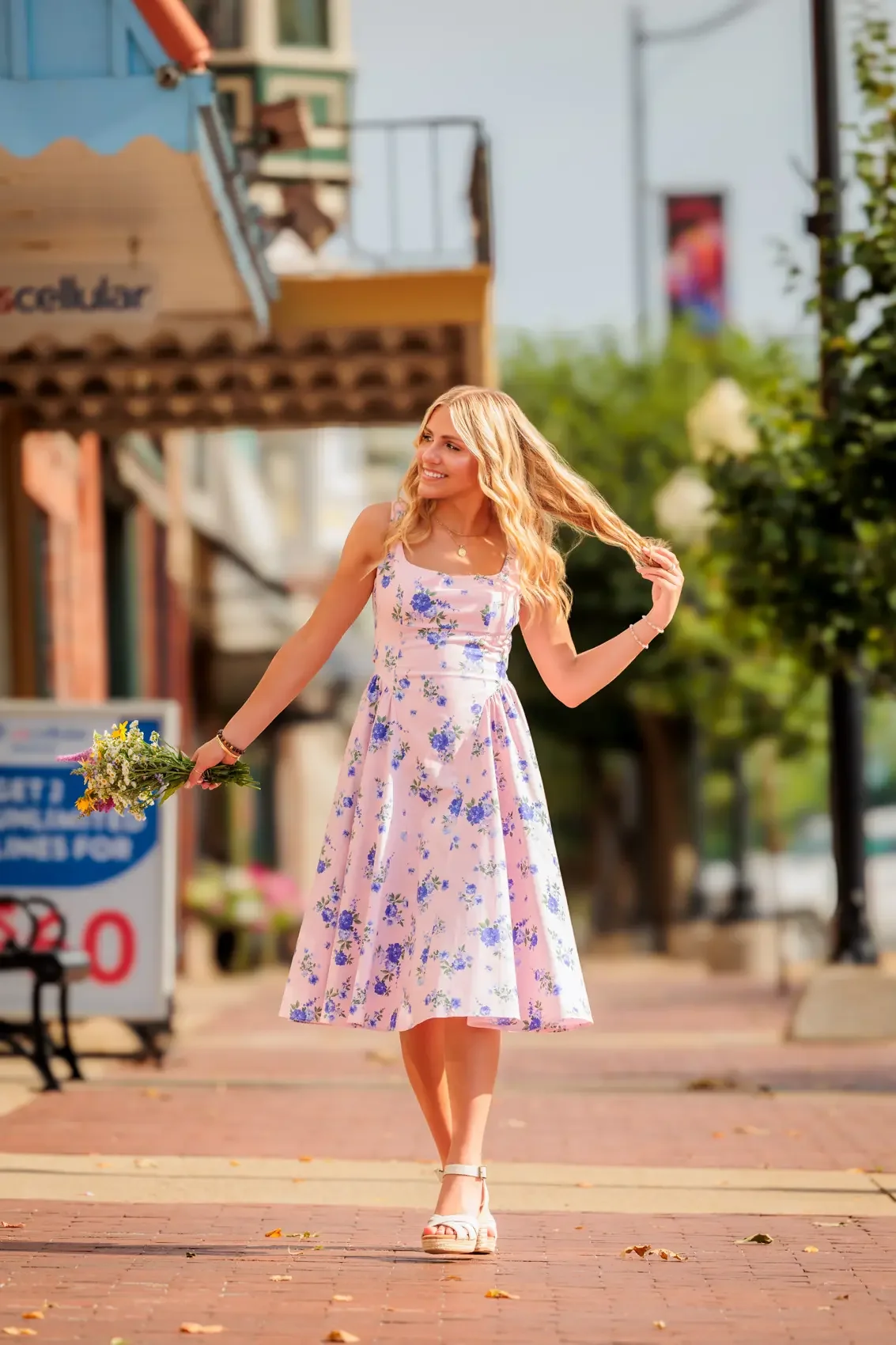Senior girl with long blonde hair wearing a pink floral dress and white sandals, holding a bouquet of flowers, walking on a brick sidewalk in a small-town street.