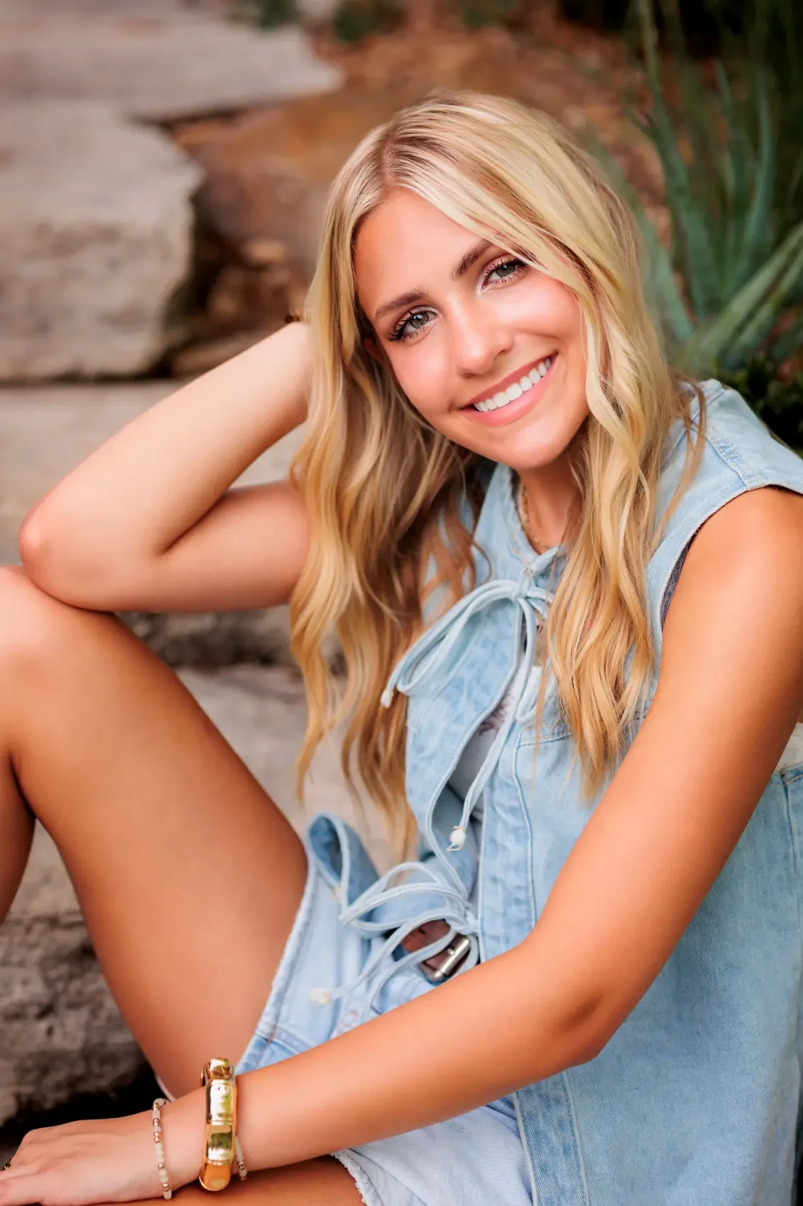 Senior girl with long blonde hair smiling and sitting outdoors, dressed in a sleeveless denim top and shorts. Photo by Des Moines senior photographer Wendy Sorensen.