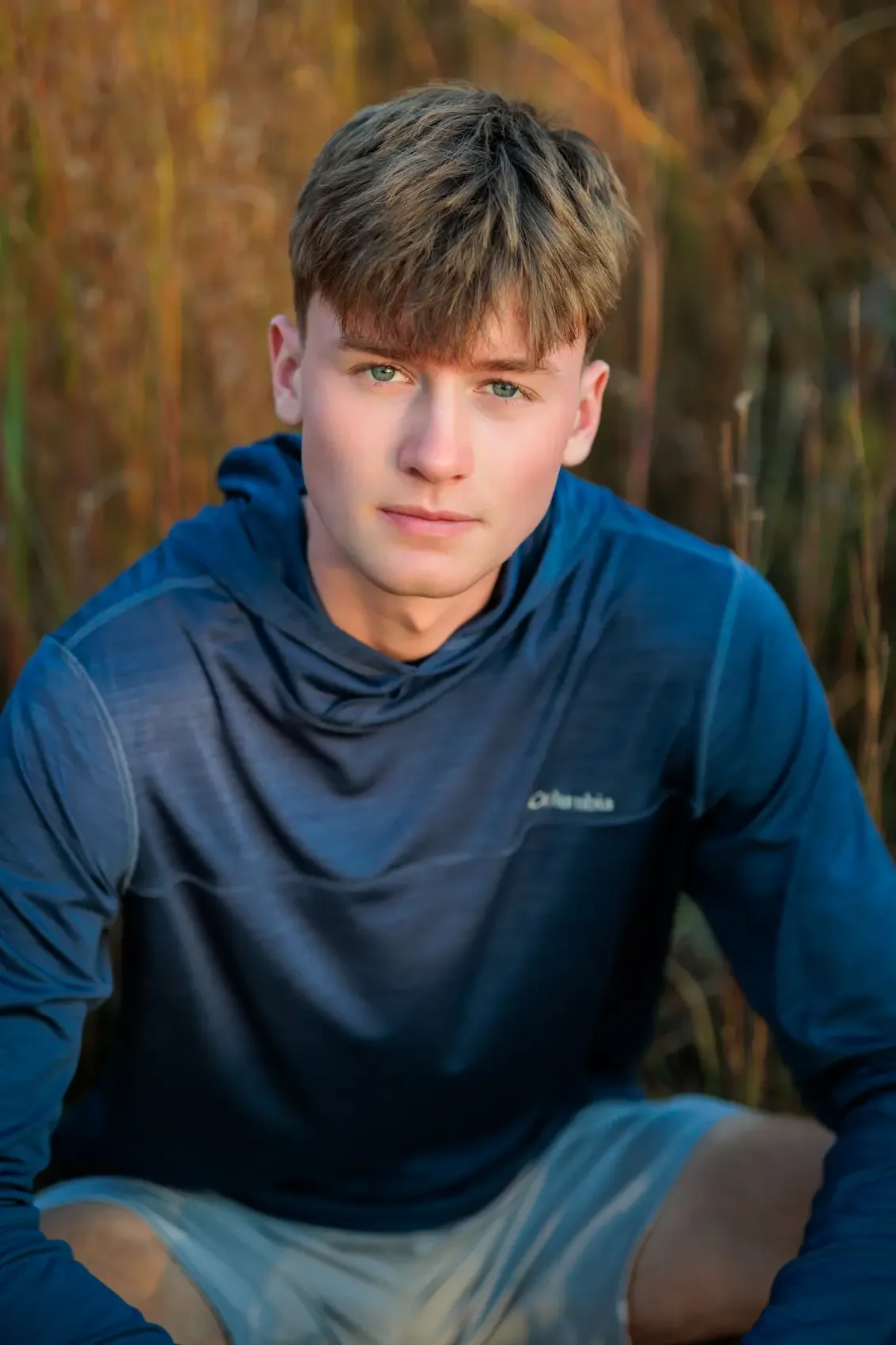 Senior boy with light brown hair and blue eyes kneeling outdoors in a natural setting, wearing a navy blue hoodie and light-colored shorts. Photo by Wendy Sorensen, a Des Moines senior photographer.