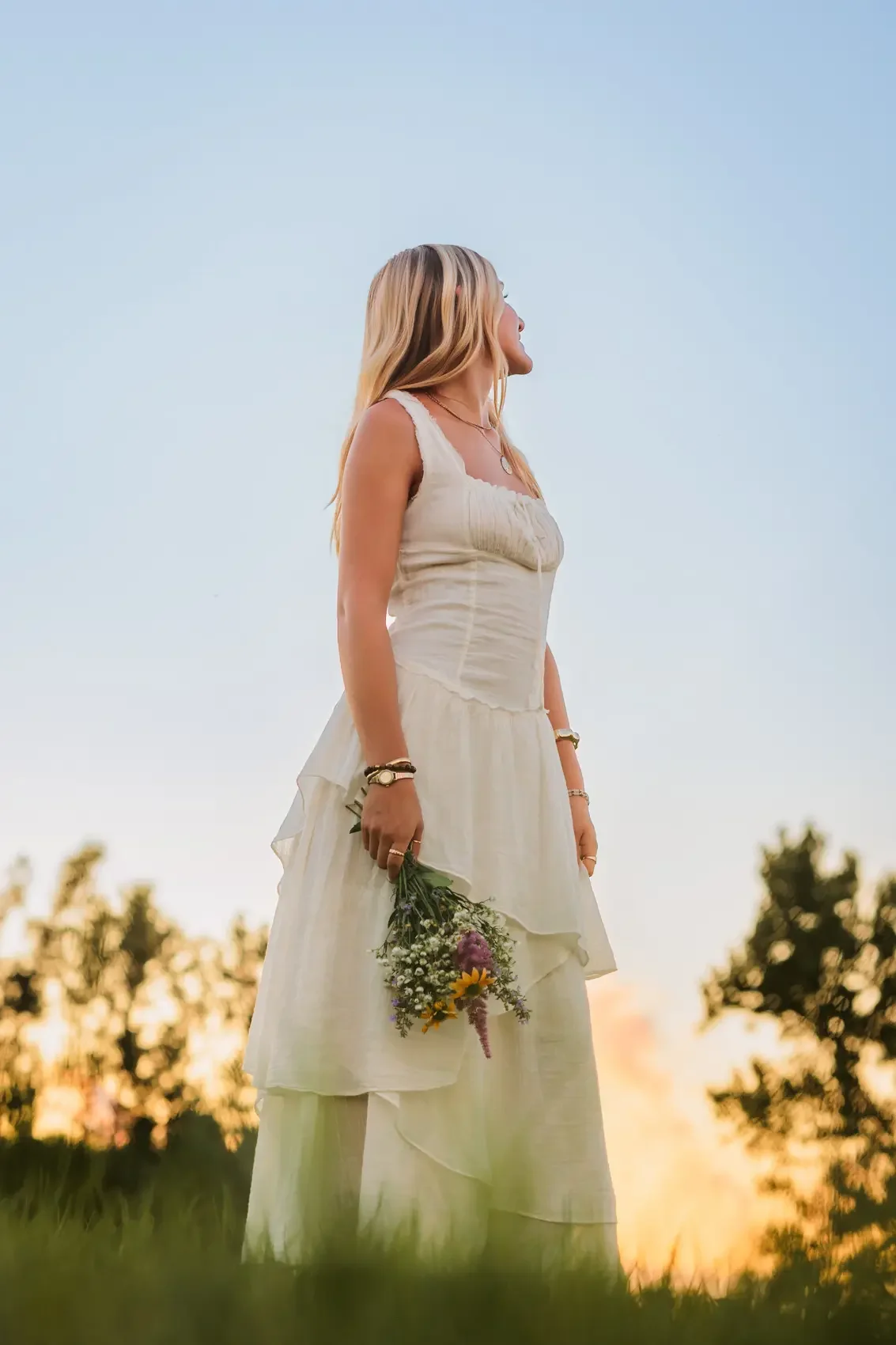 Senior girl with long blonde hair outside during sunset, holding a small bouquet of wildflowers, wearing a white bohemian dress with layered fabric and accessories.