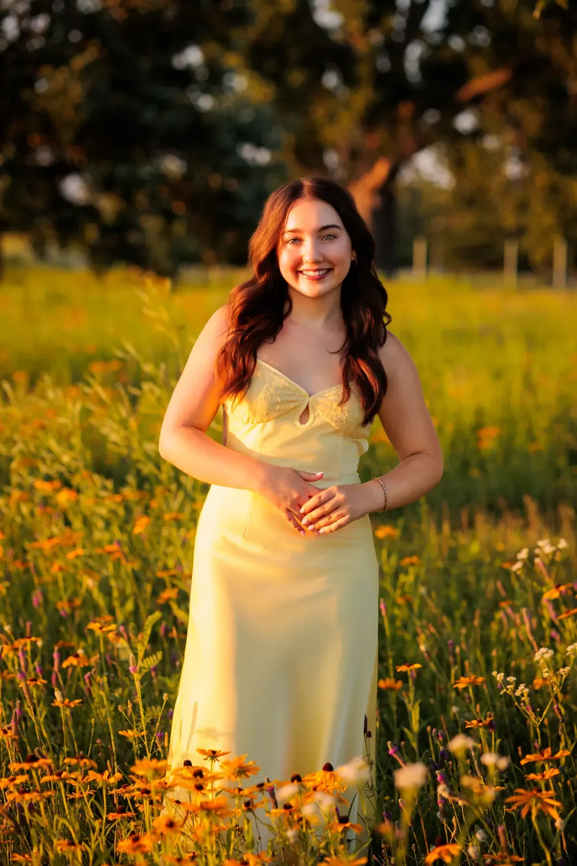 A young woman in a yellow dress standing in a field of wildflowers during sunset, smiling at the camera.