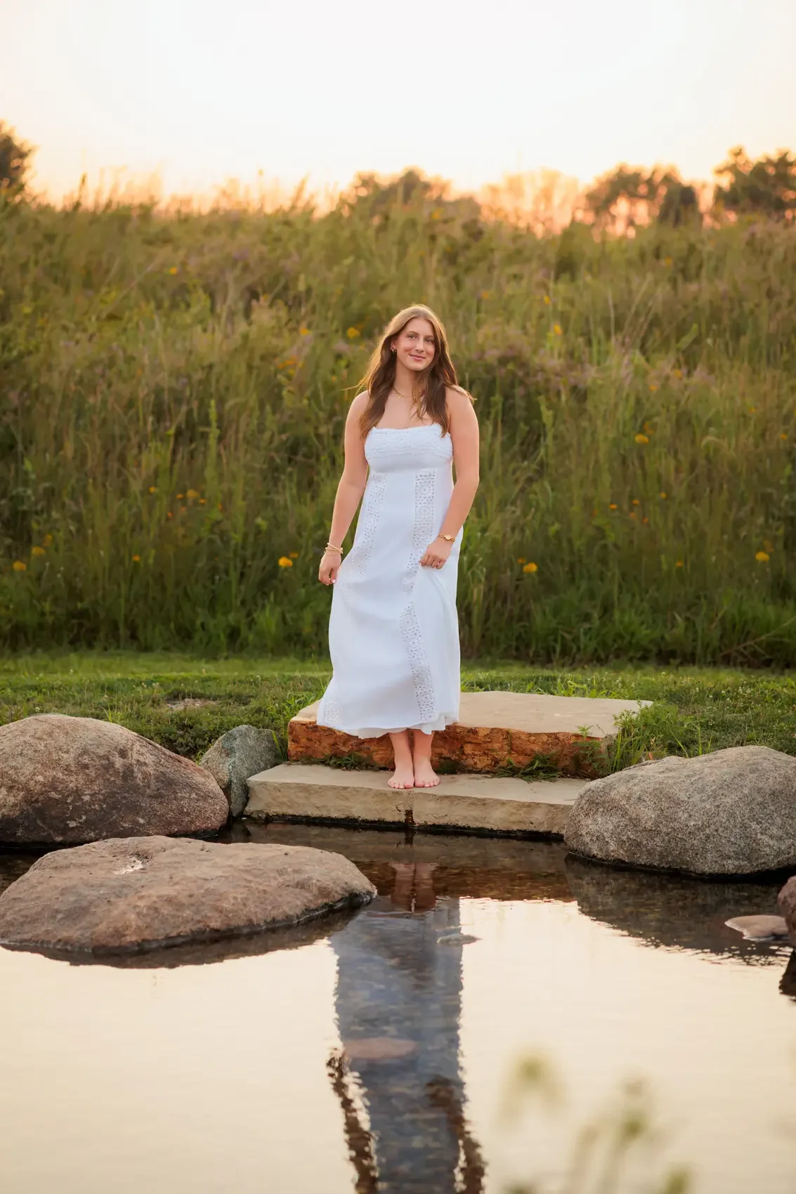 Senior girl  in a white dress standing barefoot on a stone ledge by a small pond with her reflection visible in the water, surrounded by grass and tall wildflowers at sunset in Des Moines, Iowa.