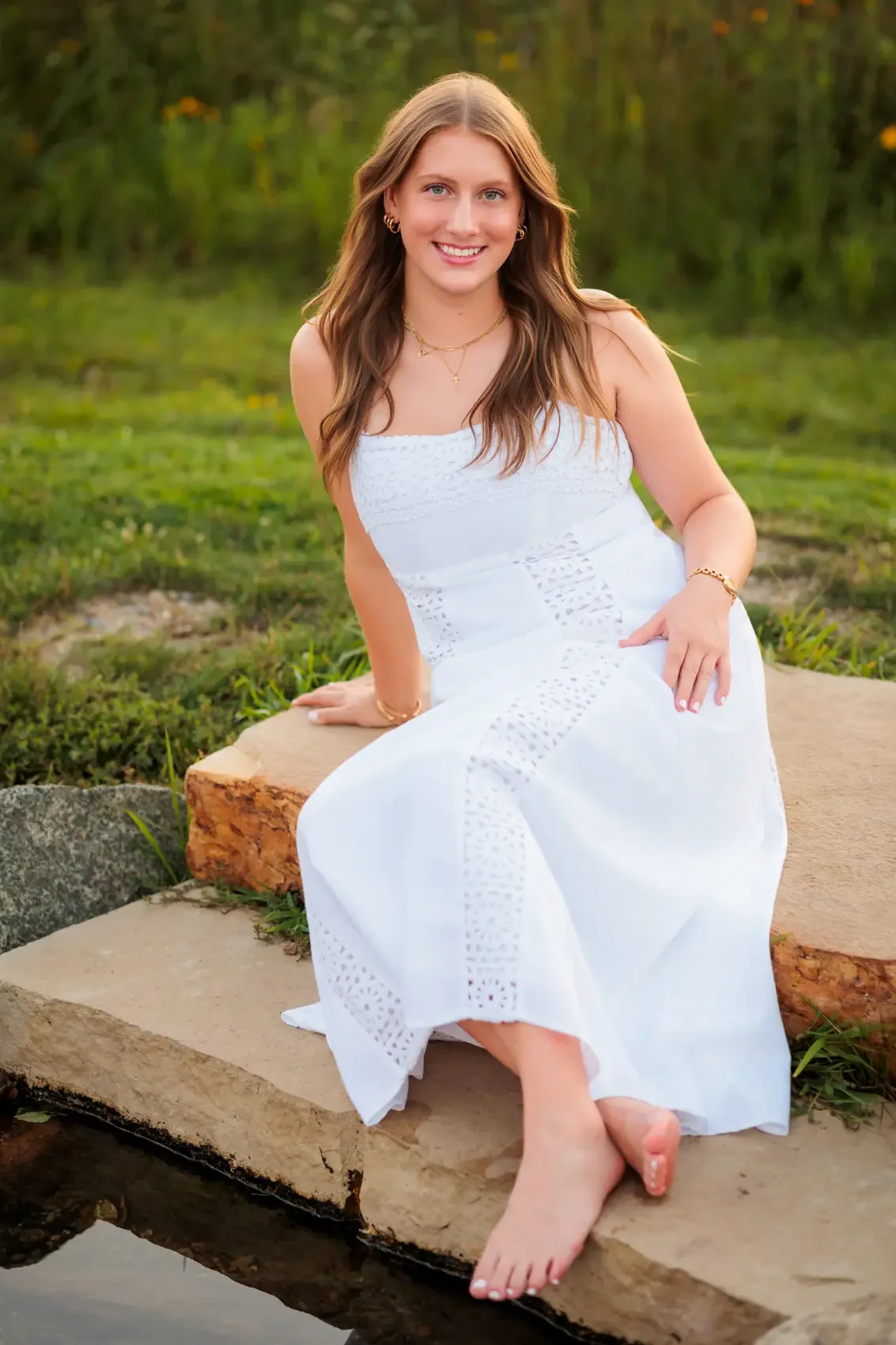 A young woman with long wavy brown hair, wearing a white sundress, sitting on a large flat rock near water, smiling at the camera in an outdoor setting surrounded by greenery.