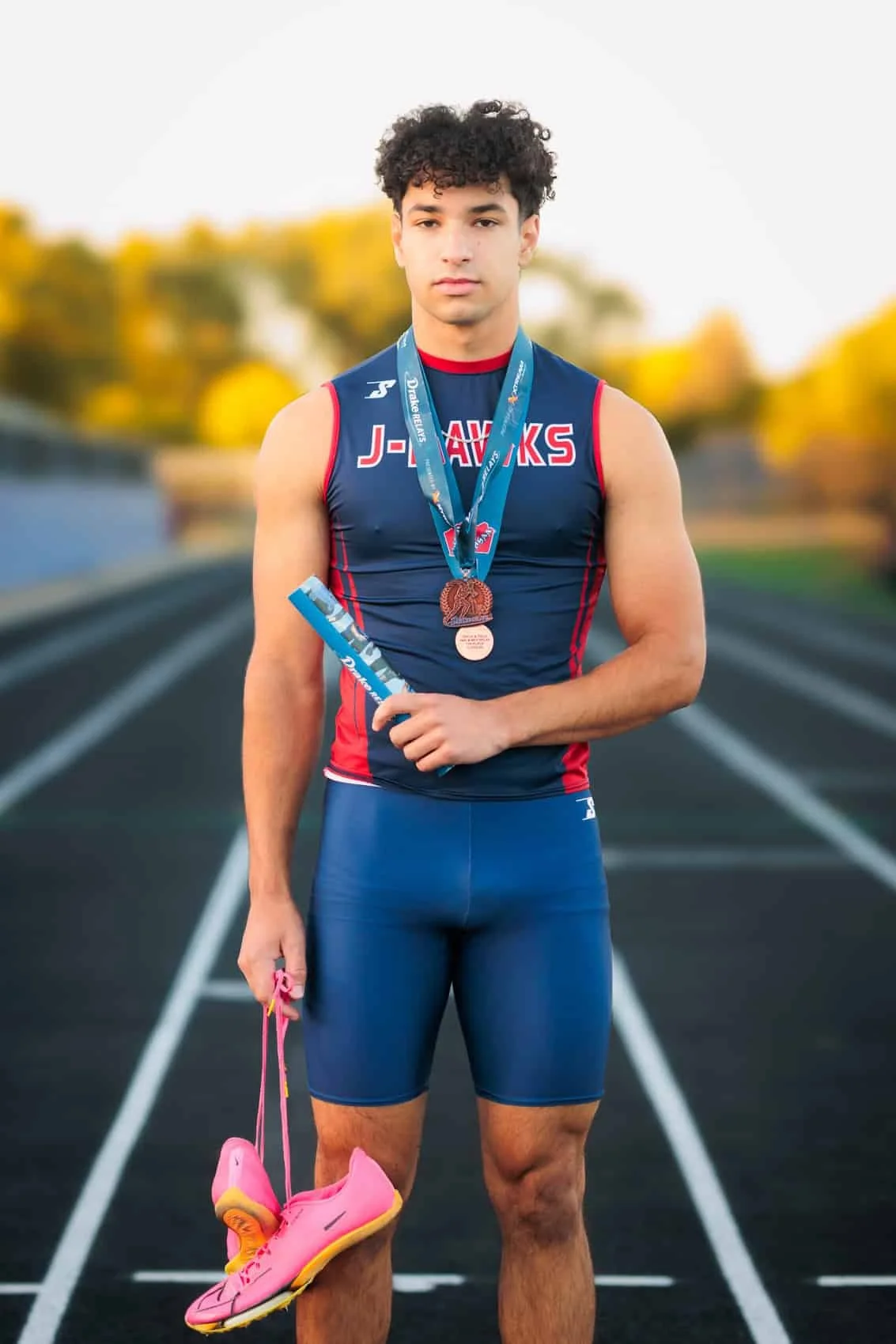 A high school senior male athlete standing on a running track outdoors, holding pink running shoes with yellow soles in his left hand, wearing a navy blue and red athletic uniform with 'J-Hawks' on the chest, and multiple medals around his neck. 