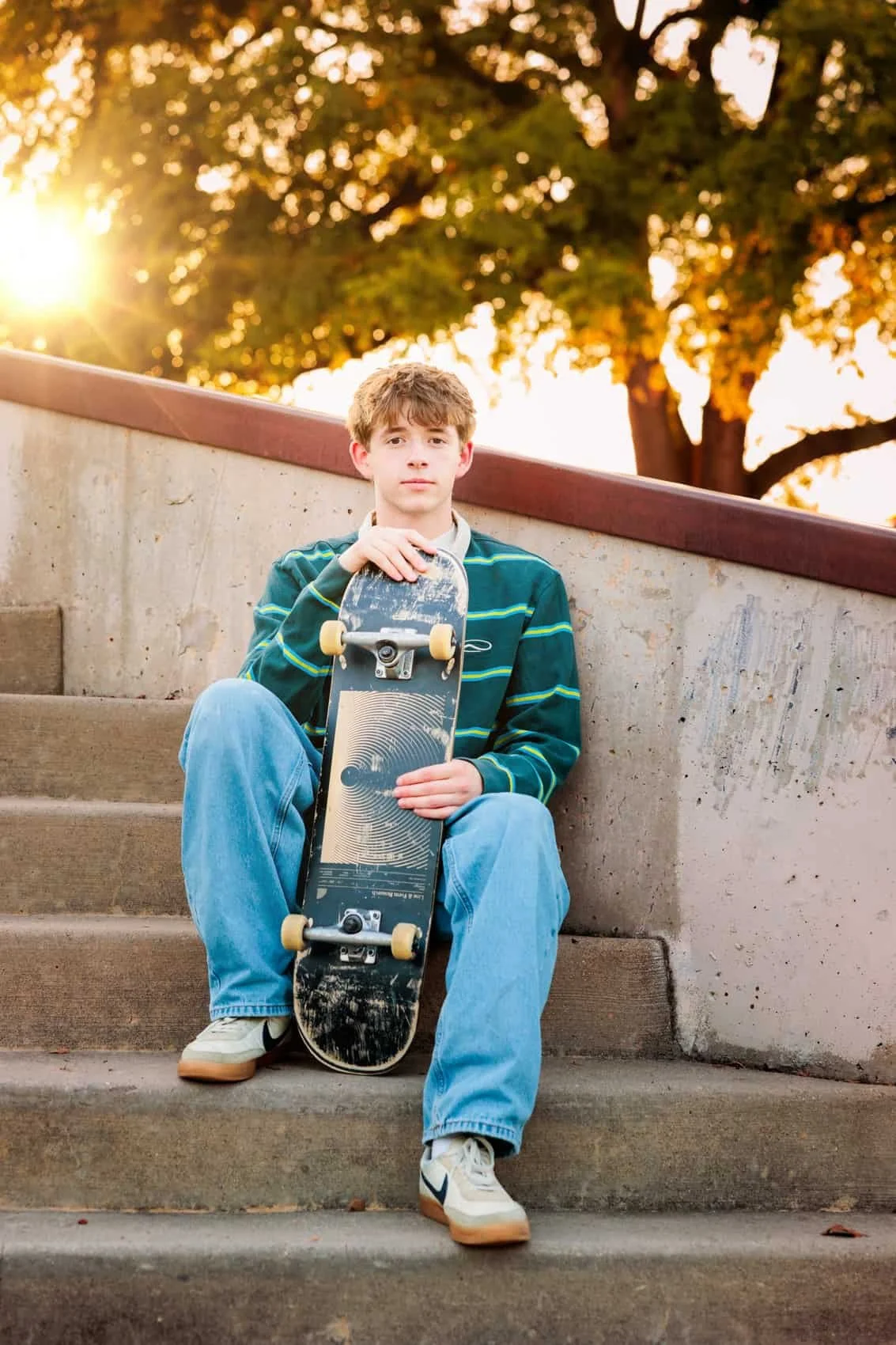  High school senior boy sitting on concrete stairs holding a skateboard, with sunlight and trees in the background during sunset.