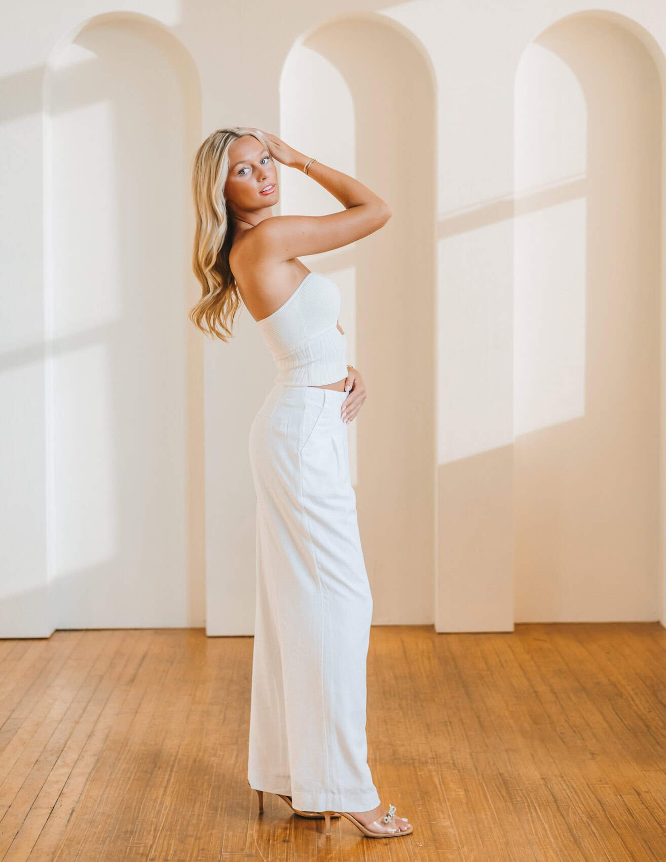 Senior girl with long blonde hair in a white strapless top and white wide-leg pants stands on a wooden floor in front of a wall with large arched windows casting shadows. Photo by Wendy Sorensen, a Des Moines senior photographer.