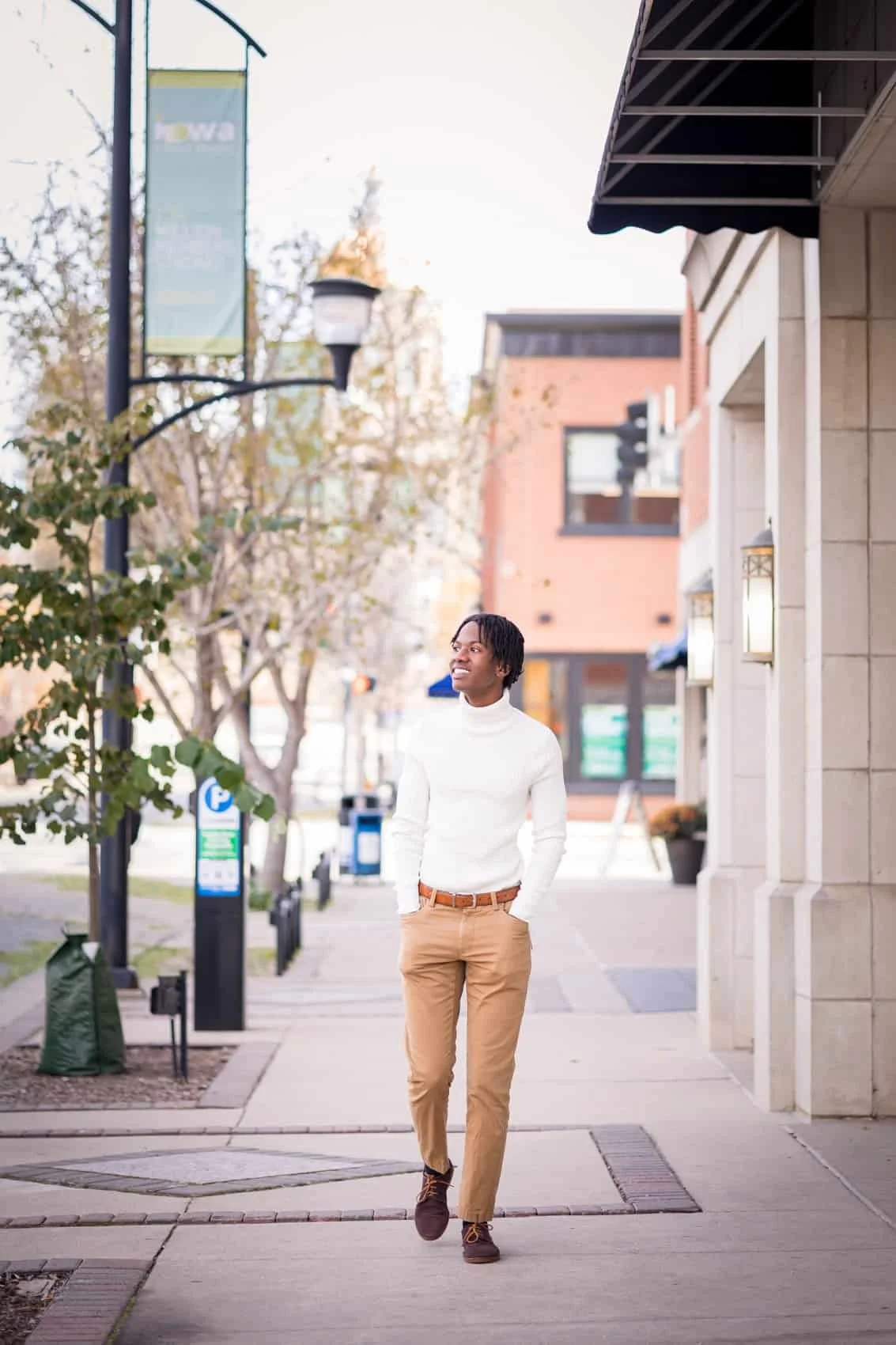  High school senior boy walking on a city sidewalk in downtown Des Moines, dressed in a white turtleneck sweater, tan pants, and brown shoes, smiling and looking to the side.