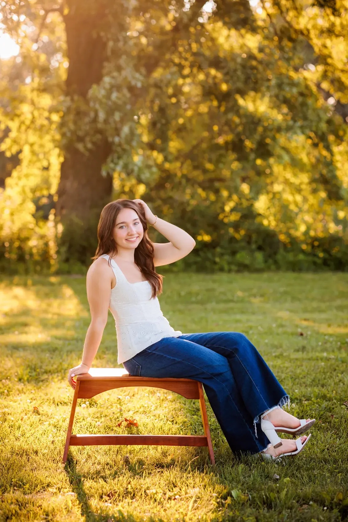 A young woman with long brown hair, wearing a white sleeveless top, blue jeans, and white high-heeled shoes, sitting on a wooden bench outdoors in a park during golden hour with sunlight filtering through trees in the background.