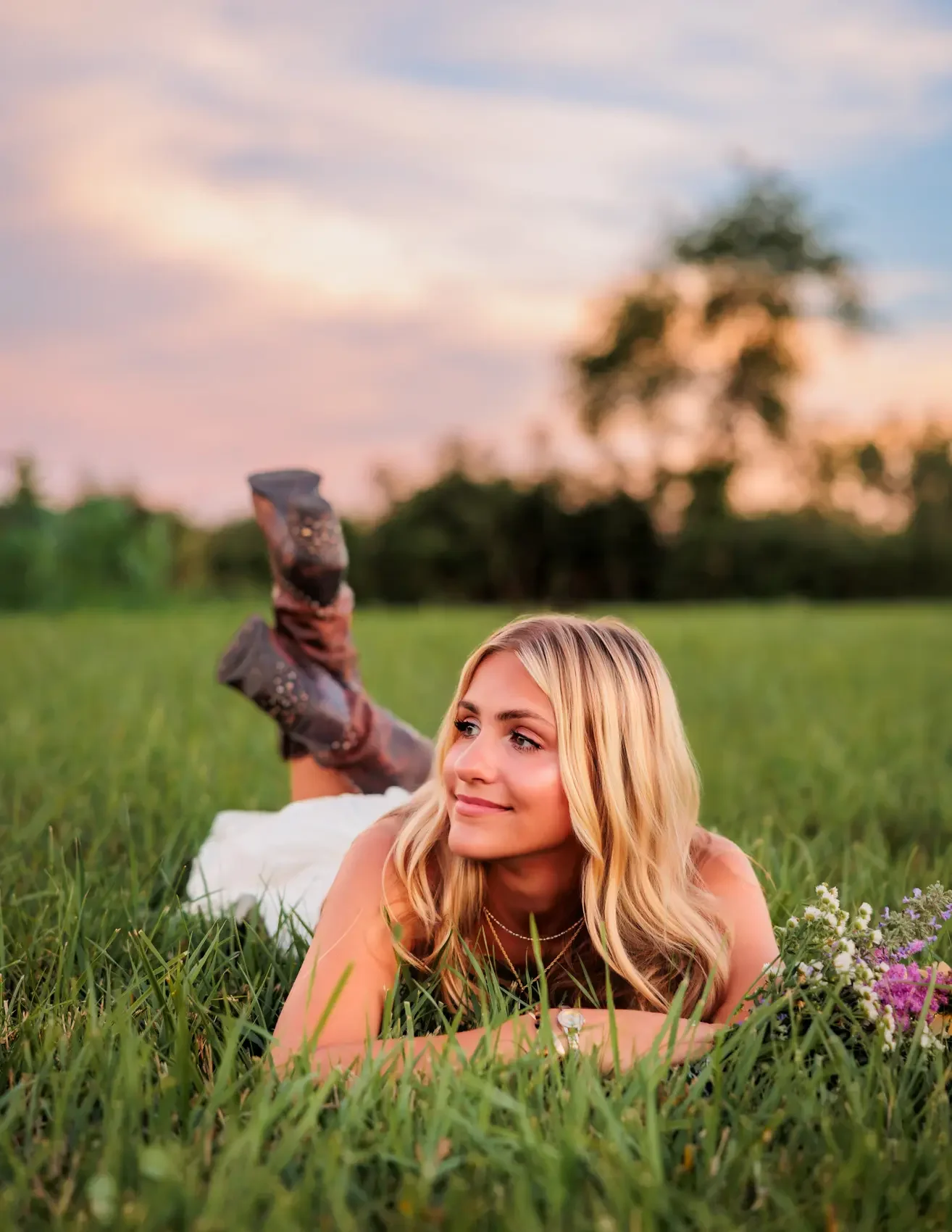 Senior girl with blonde hair lying on green grass in a field during sunset, smiling and looking to the side.