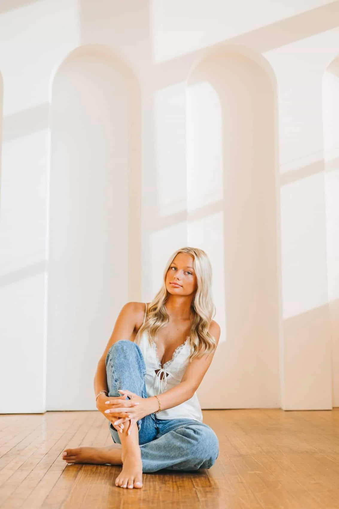 Senior girl with long blonde hair sitting on a wooden floor in a sunlit indoor photo studio in Des Moines with a white arched wall in the background.