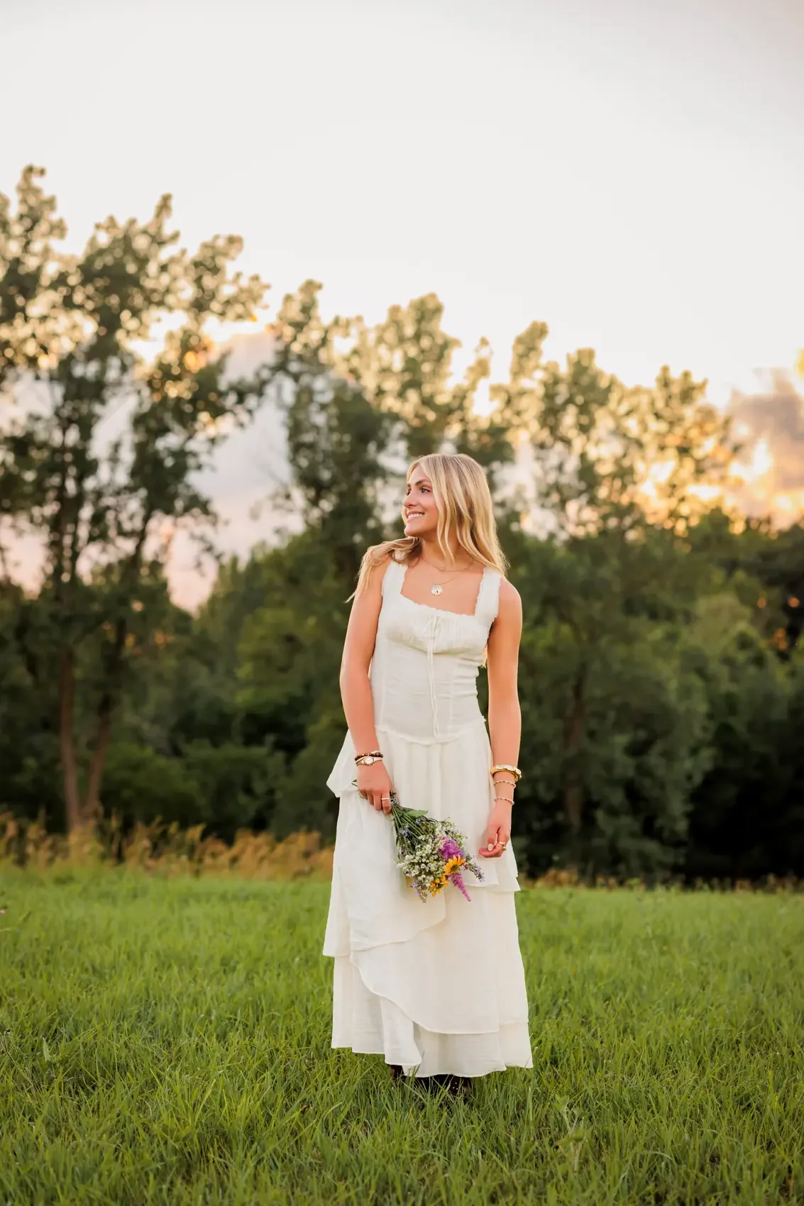 Senior girl in a white dress holding a small bouquet of flowers, standing on a grassy field with trees and a sunset sky in the background.