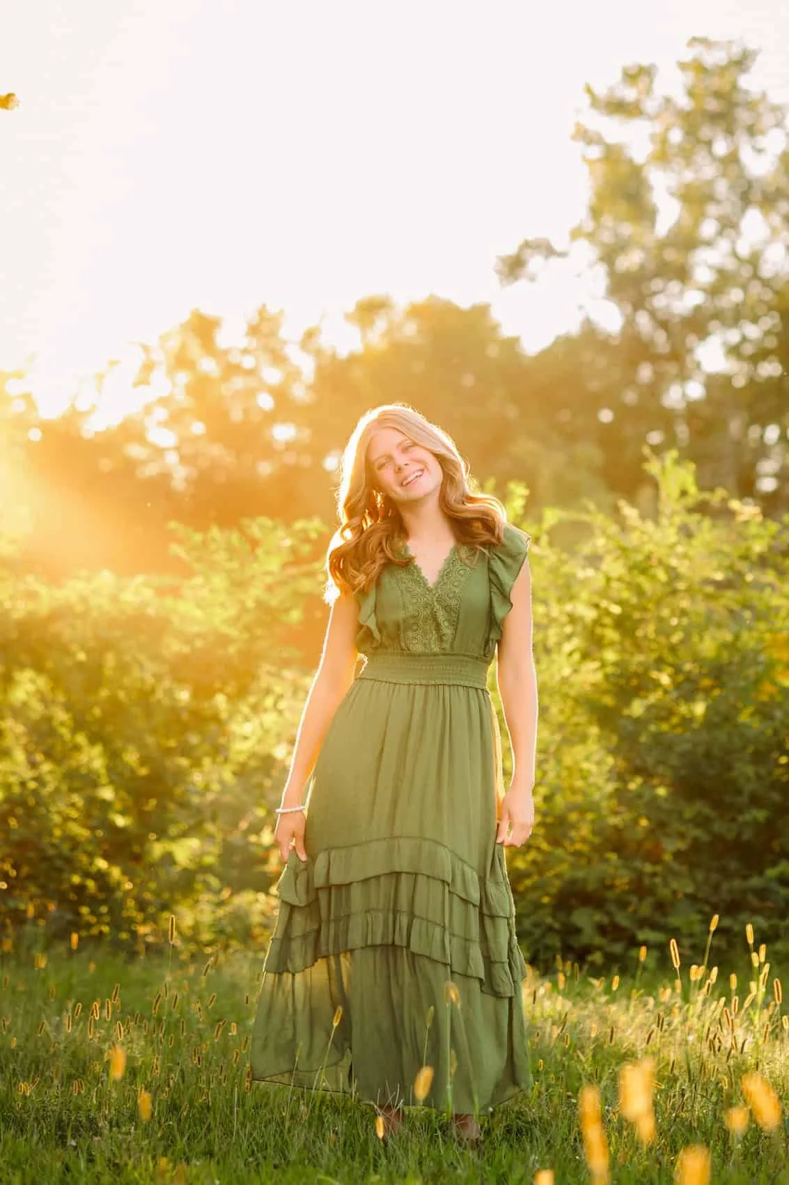  High school senior girl wearing a green dress smiling and standing in a field of grass and yellow flowers with trees in the background at Water Works Park in Des Moines during sunset.