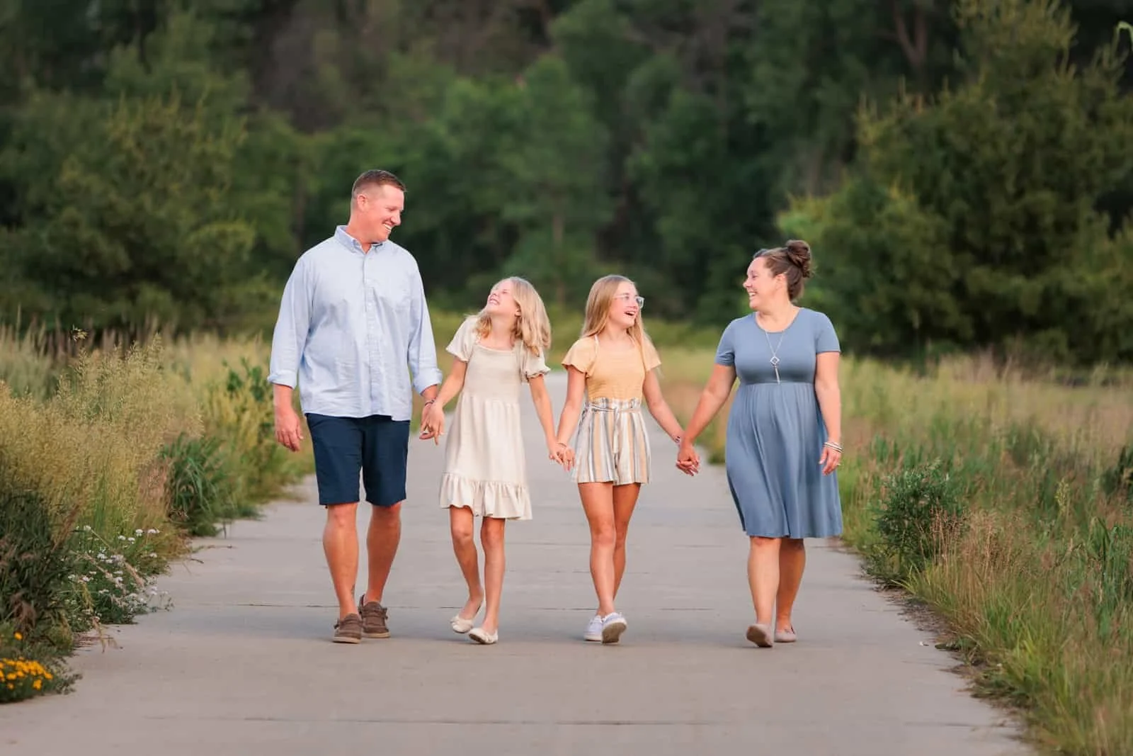 Family of 4 walking along a park pathway holding hands and laughing.