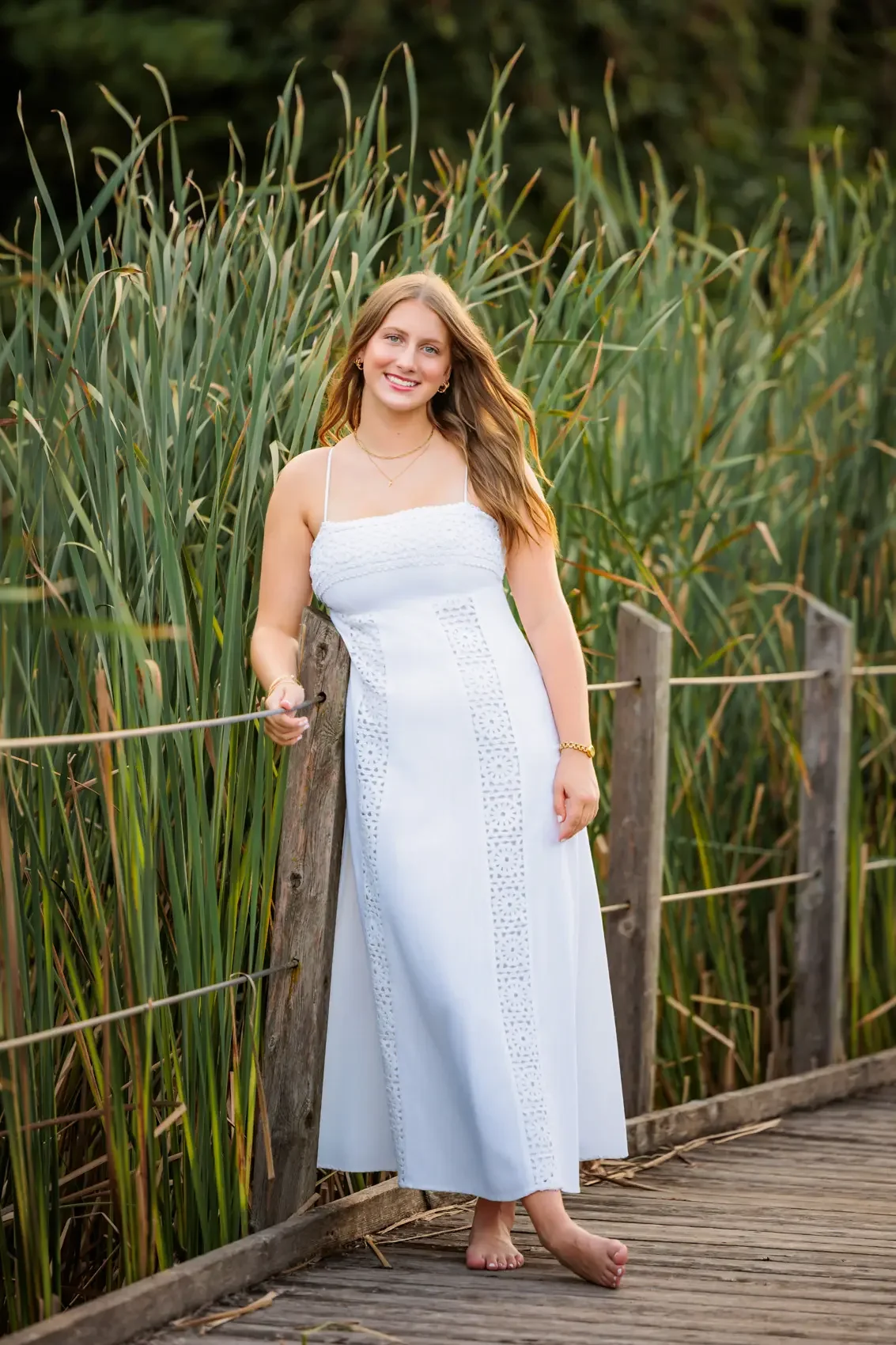 A smiling senior girl in a white dress standing barefoot on a wooden walkway next to tall green grass and reeds in Des Moines.
