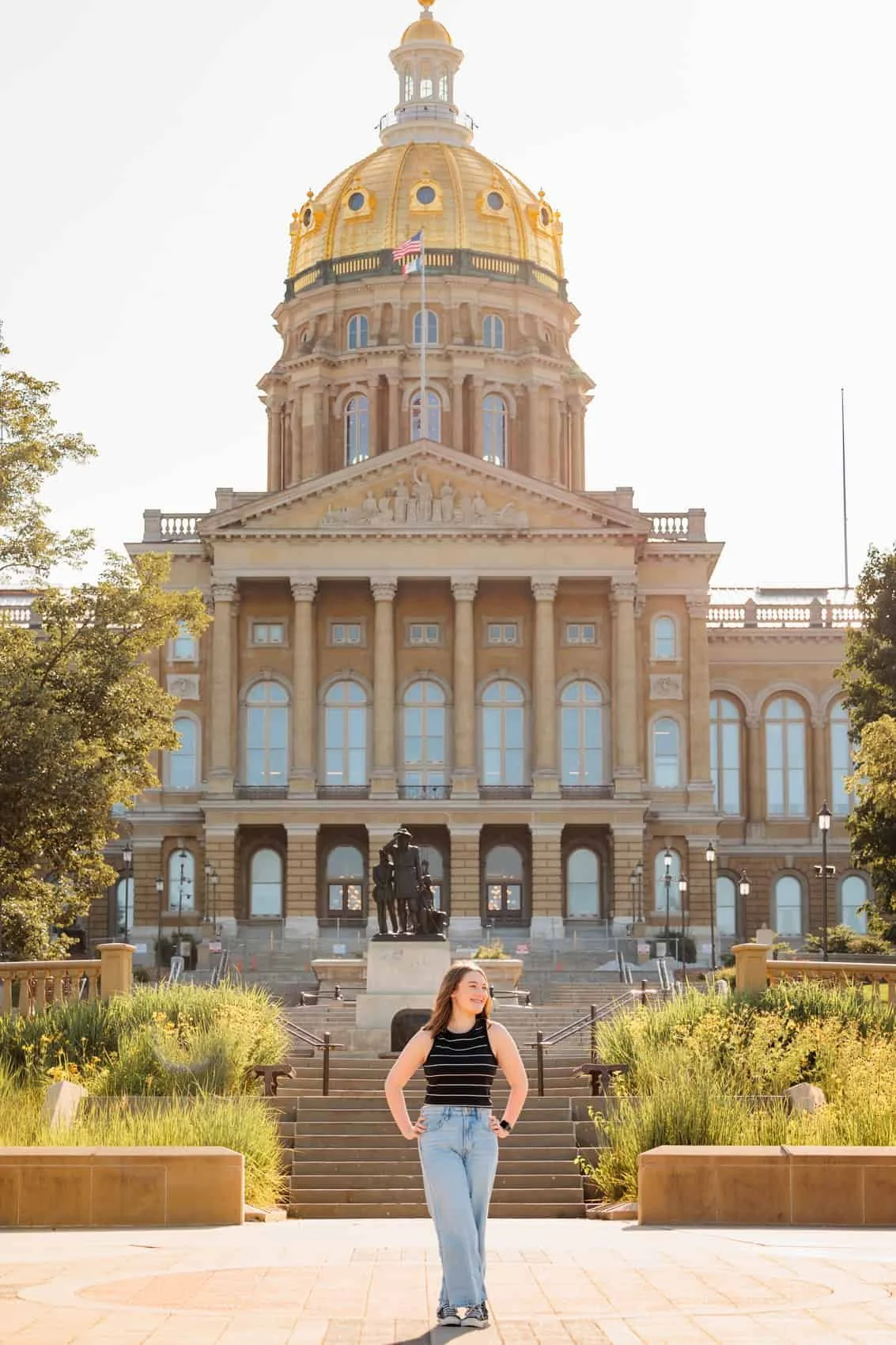  High school senior girl standing outdoors in front of the Iowa State Capitol building with a large golden dome, stairs, and a statue, surrounded by trees and greenery.