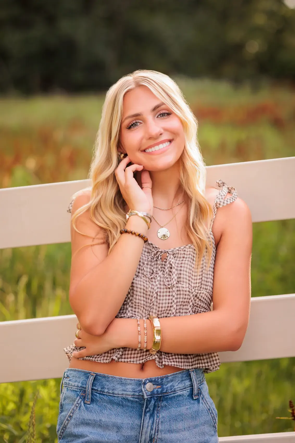 Senior girl with long blonde hair smiling outdoors, wearing a checkered sleeveless top and blue jeans, standing in front of a white fence and greenery. Photo by Des Moines senior photographer Wendy Sorensen.