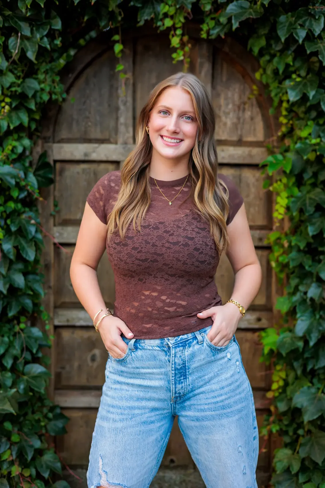 Senior girl with long wavy hair, wearing a brown lace top and light blue ripped jeans, standing outdoors in front of a wooden gate surrounded by green ivy, smiling at the camera. Photo by Wendy Sorensen, a Des Moines senior photographer.