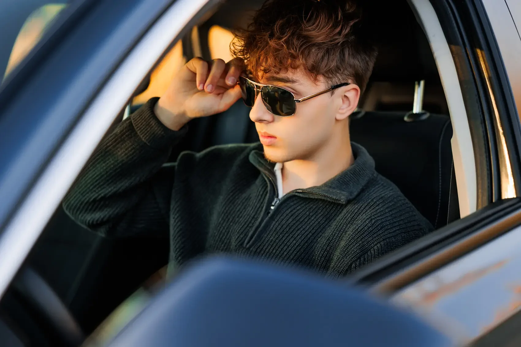Young man with curly hair wearing sunglasses and a black zip-up sweater sitting in a car.