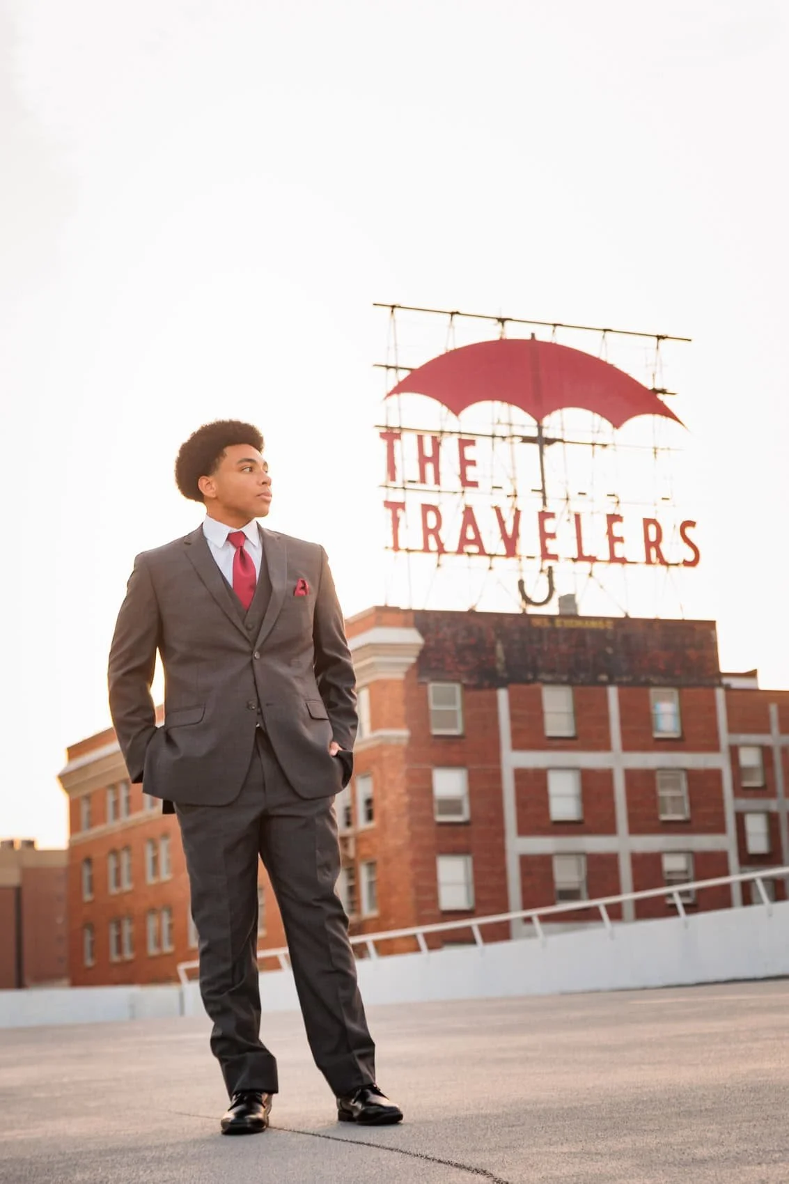 Senior boy in a gray suit and red tie standing with hands in pockets on a parking garage rooftop during sunset, with a sign that reads 'The Travelers' and has a red umbrella logo in the background, in downtown Des Moines, Iowa.