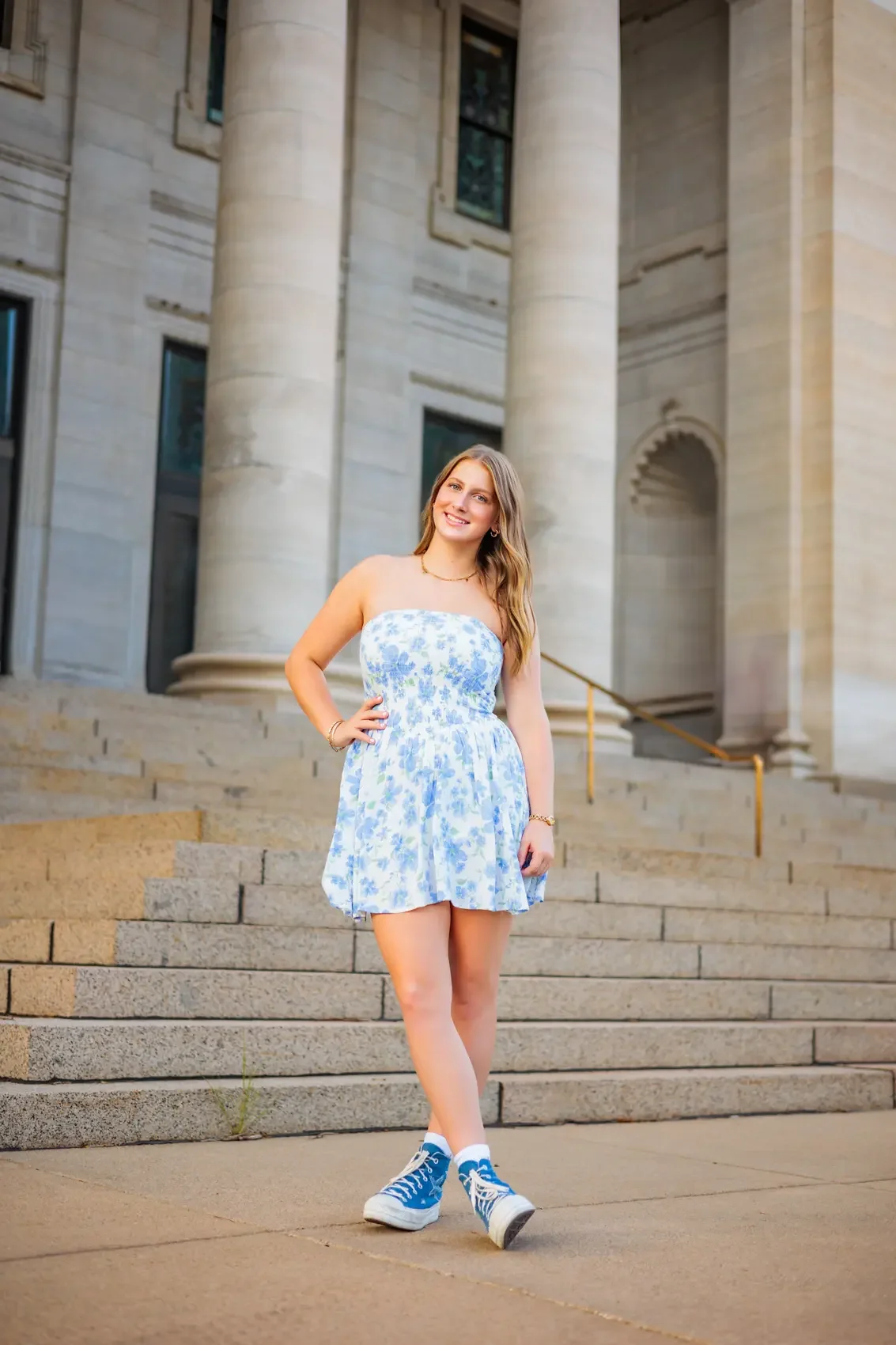 Senior girl standing on steps in front of a building with columns. She is smiling, wearing a white and blue floral strapless dress and blue sneakers, posing for her senior picture session in Des Moines.