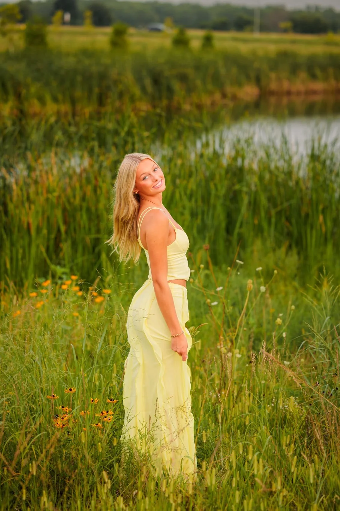 Senior girl in a yellow crop top and yellow flowy pants standing in tall grass near water, smiling at the camera.