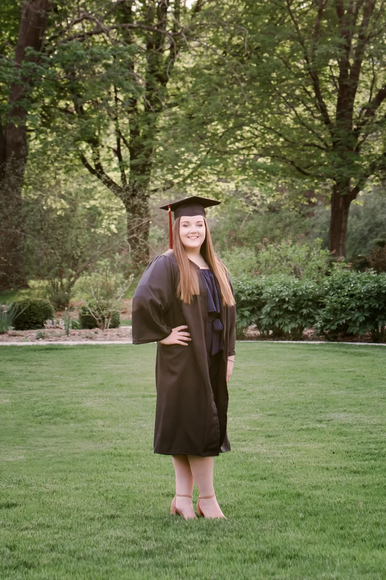 College senior woman with long brown hair wearing cap and gown in a green, nature area on the campus of Iowa State University.