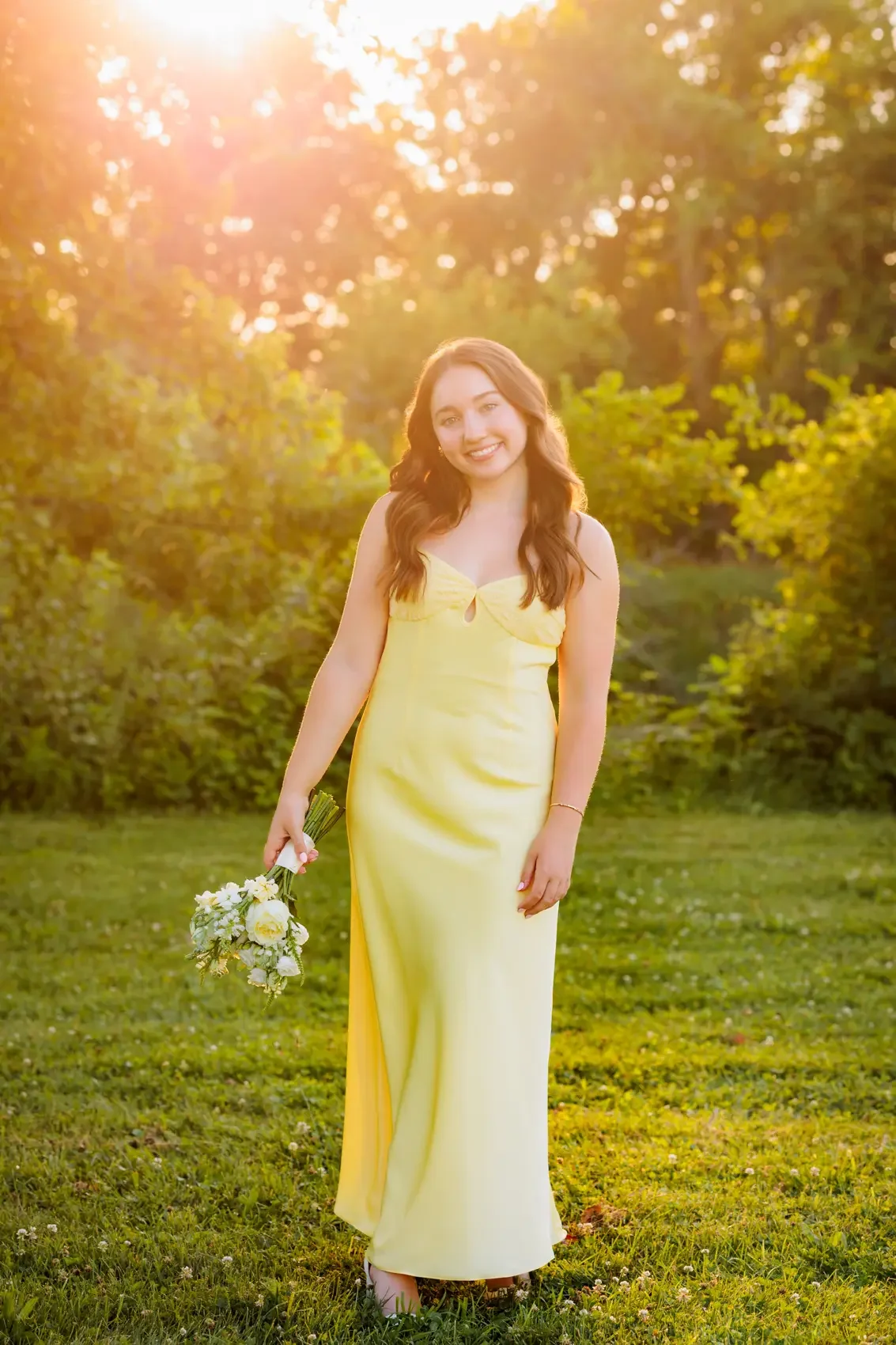High school senior girl in a yellow dress holding a bouquet of flowers, smiling outdoors during sunset with trees in the background.