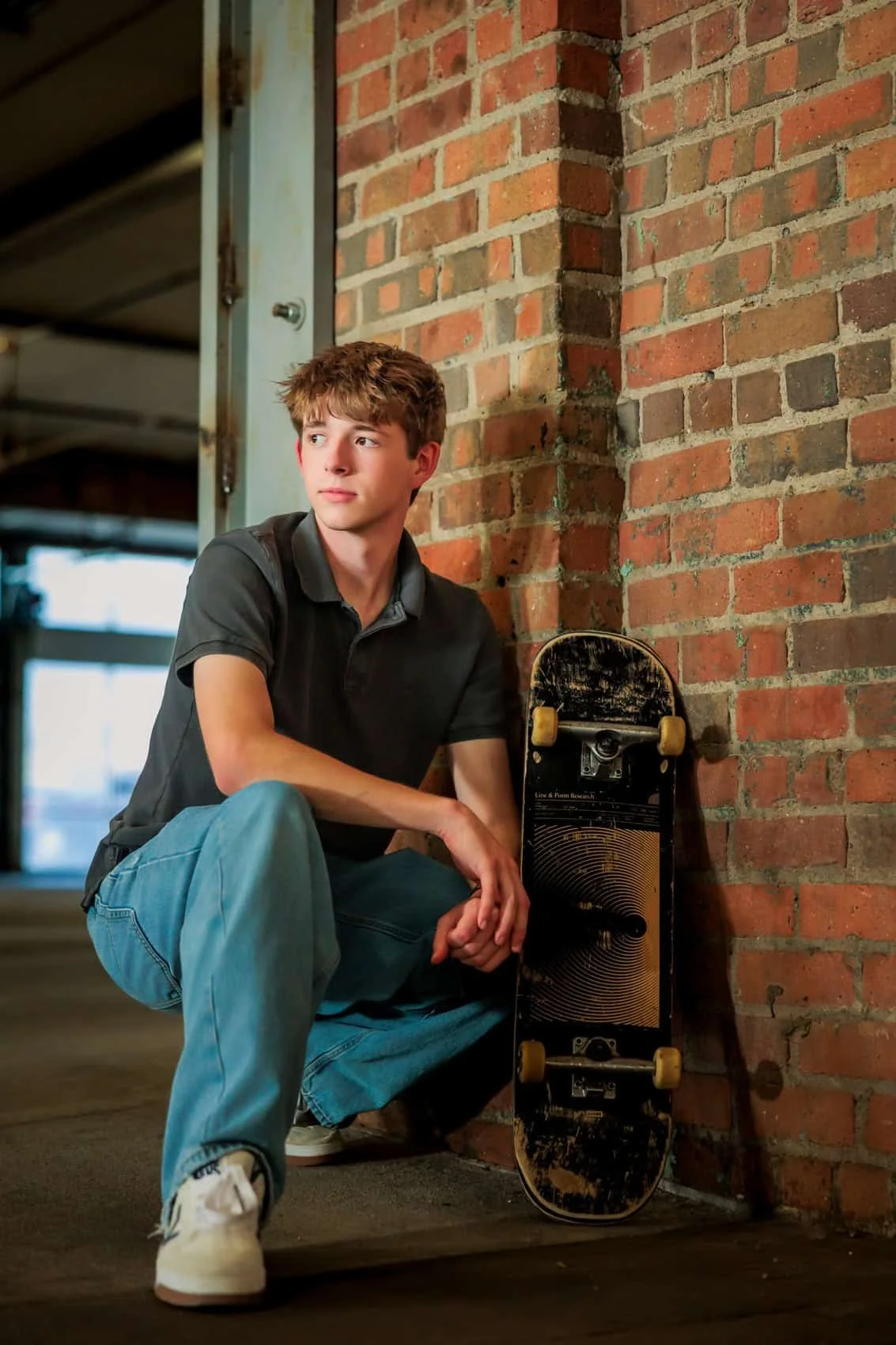  High school senior boy with brown hair, wearing a black polo shirt and light blue jeans, squatting next to a skateboard with a worn black top and yellow wheels, leaning against a brick wall in an indoor setting with natural light.