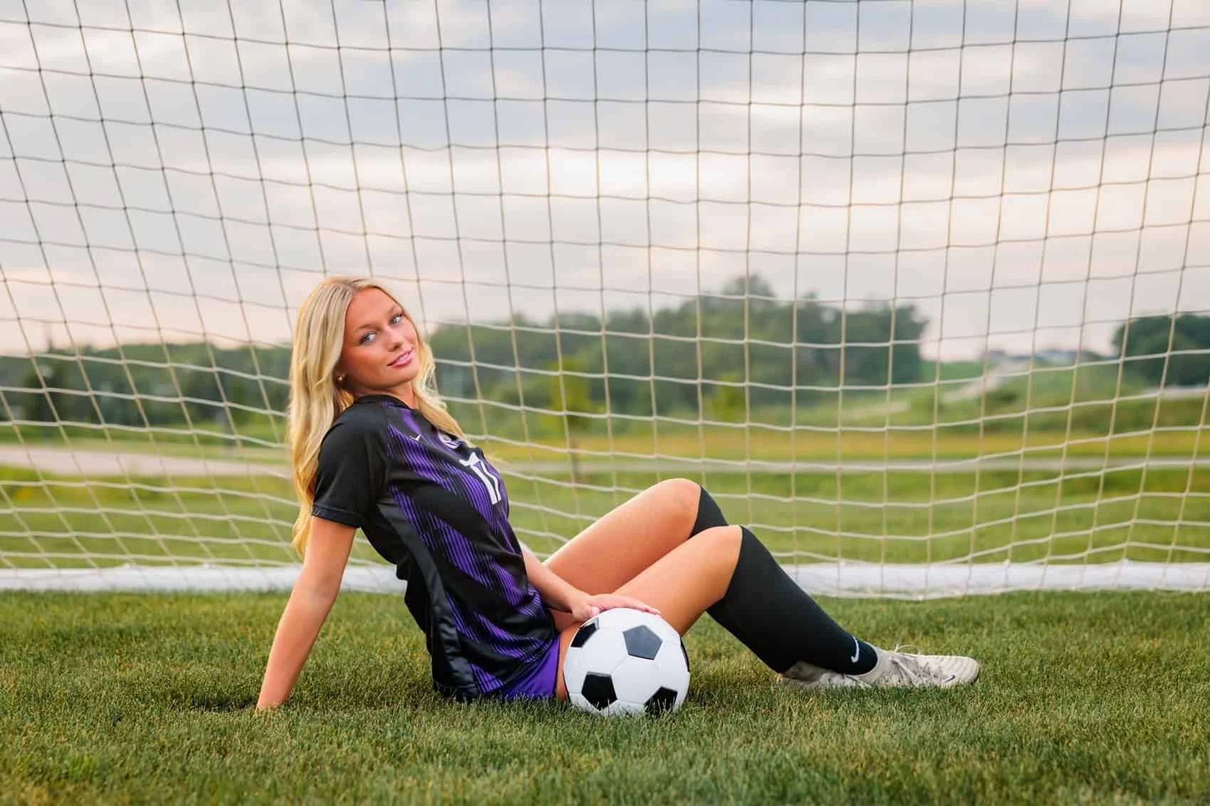 Senior girl in soccer uniform sitting on grass in front of goal post with soccer ball