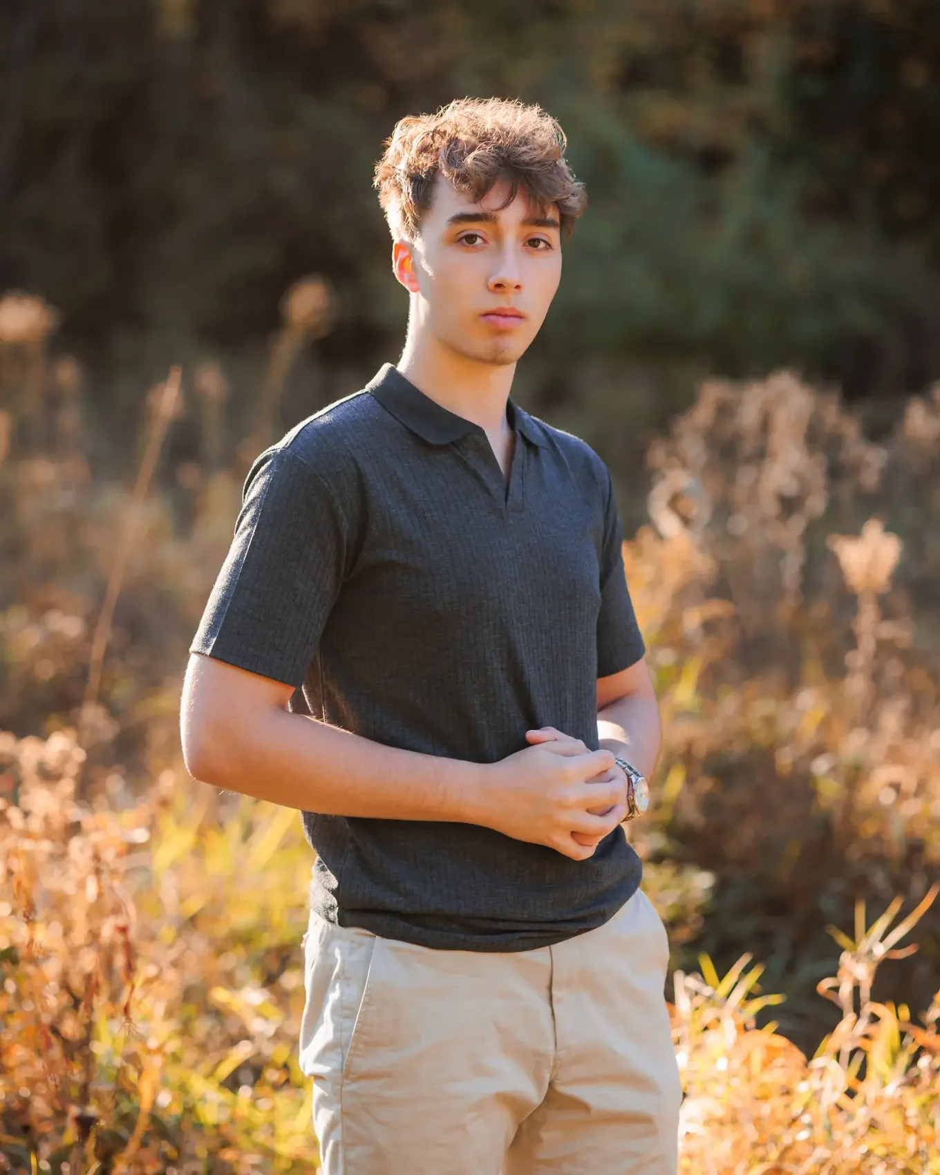Senior boy standing outdoors in a field with dry plants and trees, wearing a dark polo shirt and khaki shorts, looking at the camera with a neutral expression.