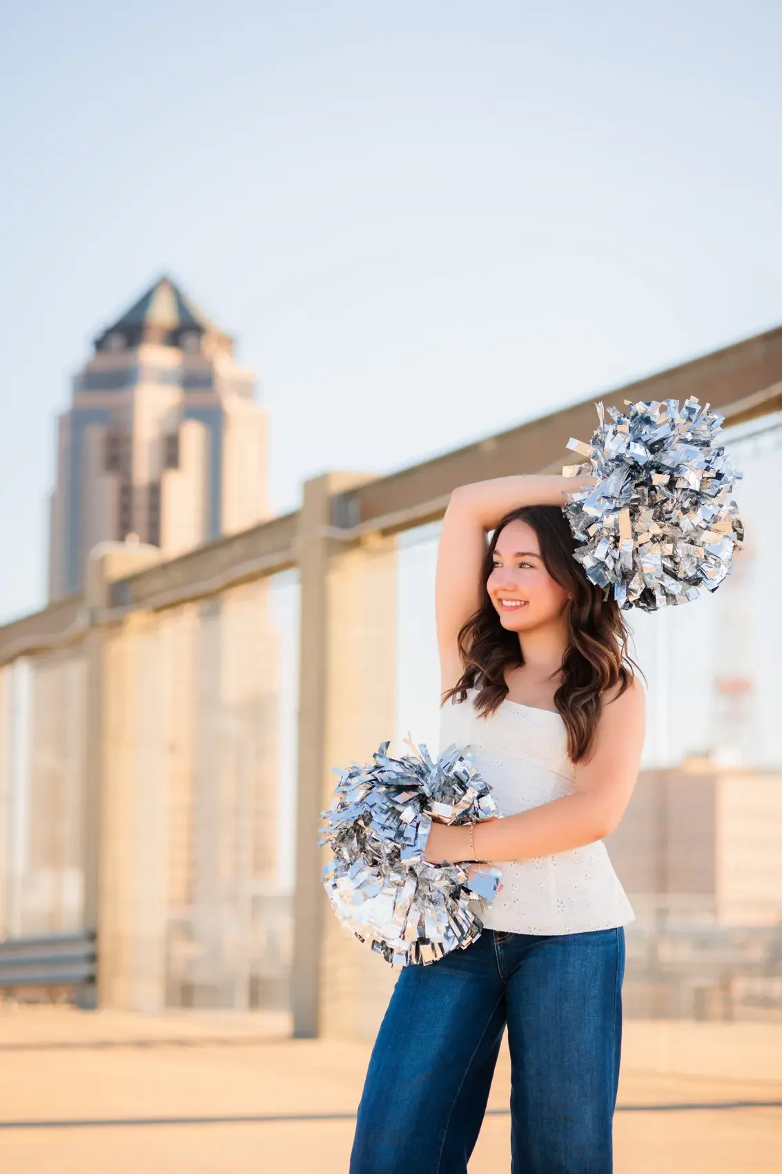 Senior girl with silver pom-poms outdoors on a parking garage rooftop in Des Moines, smiling and wearing a white top and blue jeans, with a city skyline in the background. Photo by Des Moines senior photographer Wendy Sorensen.