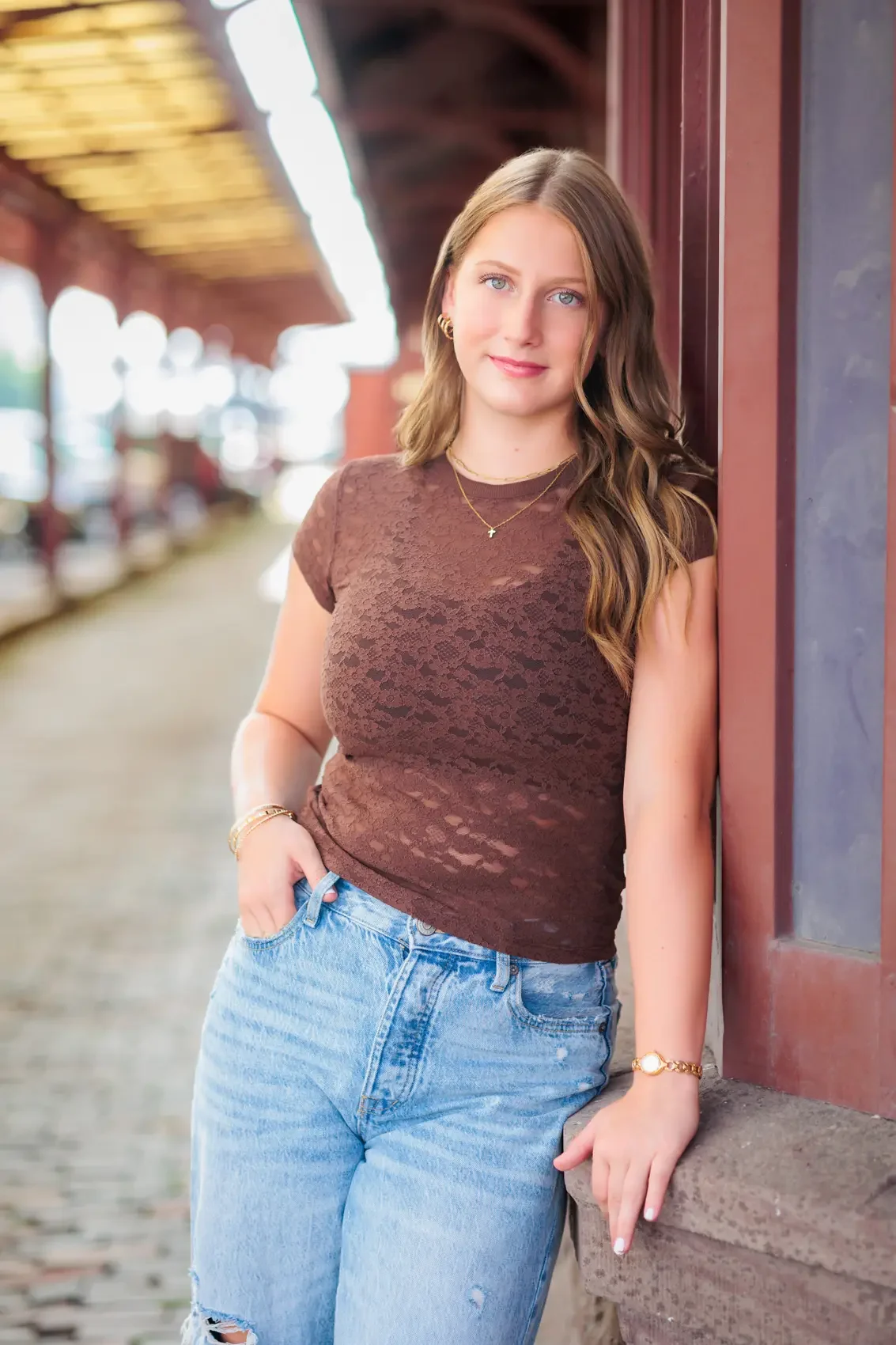 High school senior girl with long brown wavy hair and blue eyes, wearing a brown lace top and light blue jeans, leans against a brick wall in an outdoor setting with a blurred train platform background.
