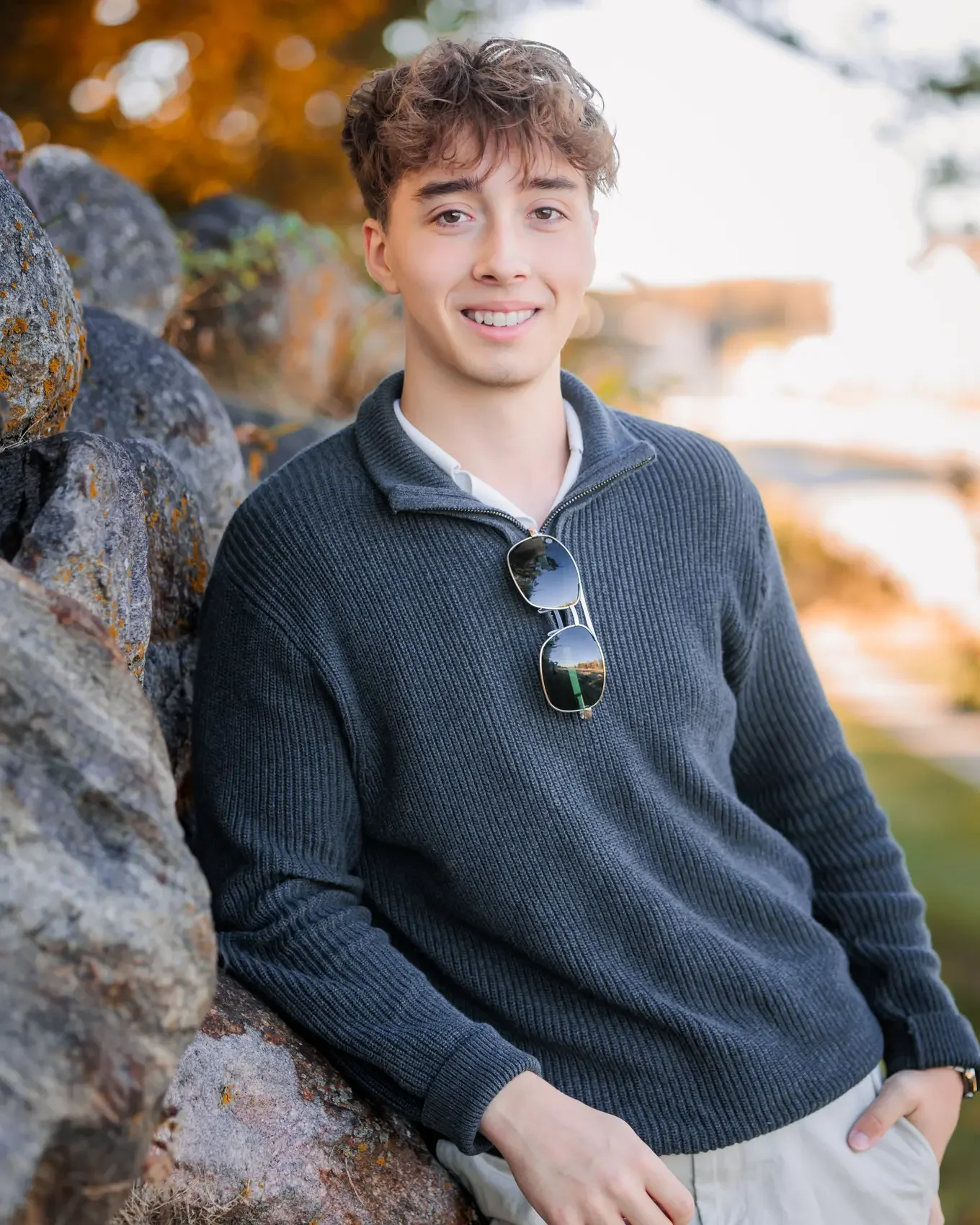 Boy with curly brown hair and fair skin smiling for senior pictures, wearing a dark blue sweater with sunglasses hanging from the collar, standing outdoors near a stone wall during golden hour in Des Moines.