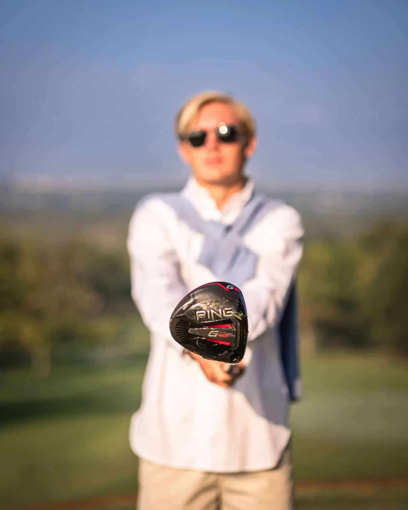  High school senior guy holding a black Ping G425 golf driver, standing outdoors at Hyperion Field Club with a blurred background of trees and sky.
