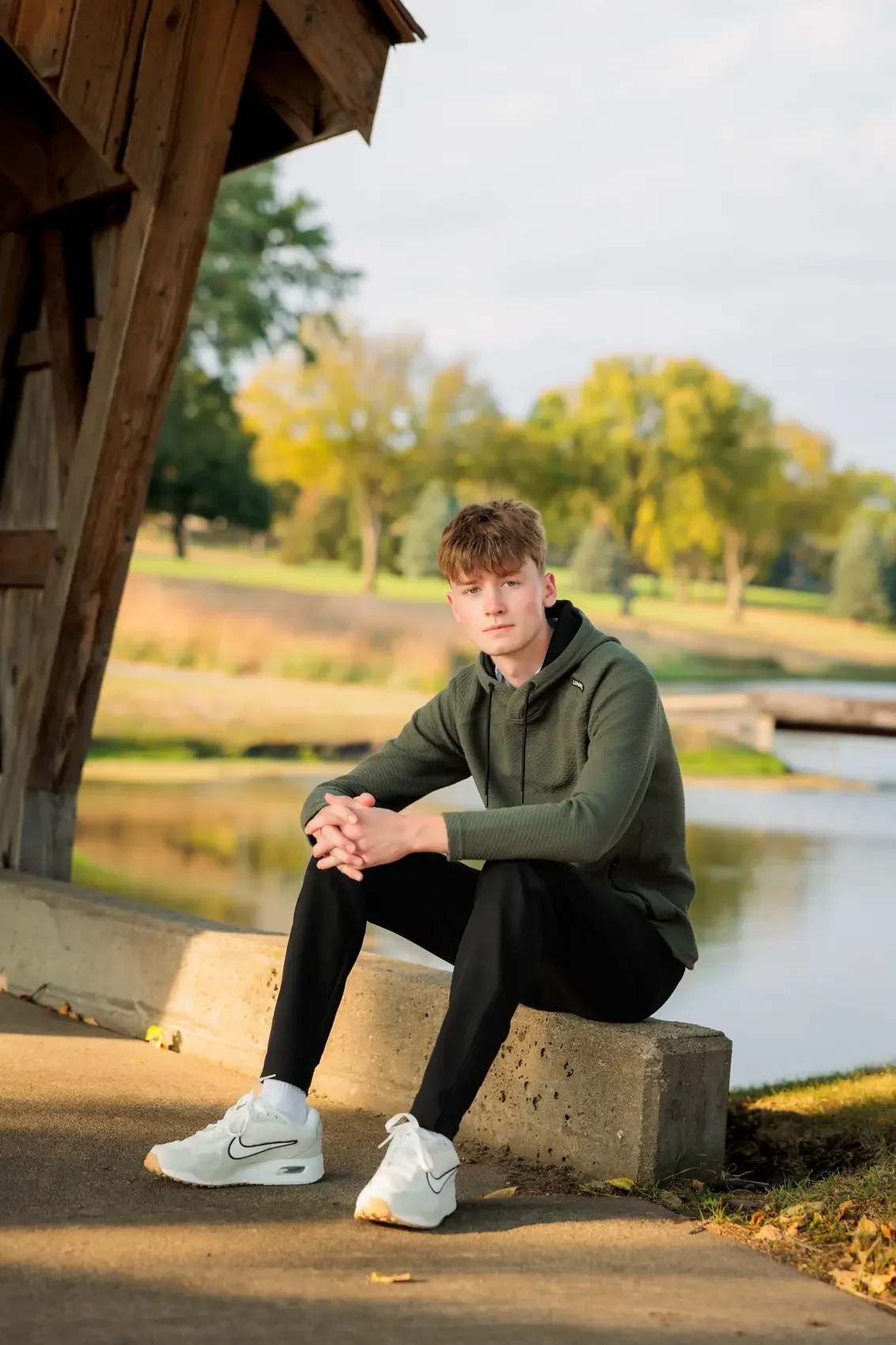 Senior boy sitting on a concrete bench outdoors near a body of water at Des Moines Golf & Country Club, wearing a dark green hoodie, black pants, and white sneakers, with a background of trees and a partly cloudy sky.