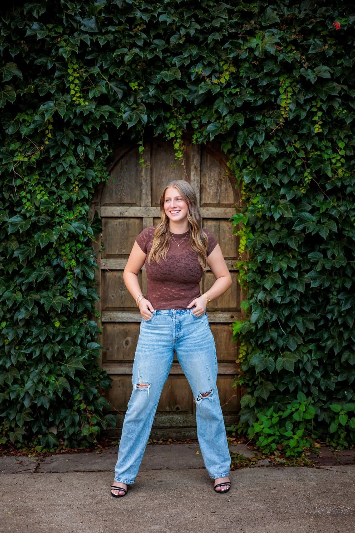 A young woman smiling and standing outdoors in front of a wooden gate surrounded by green ivy.
