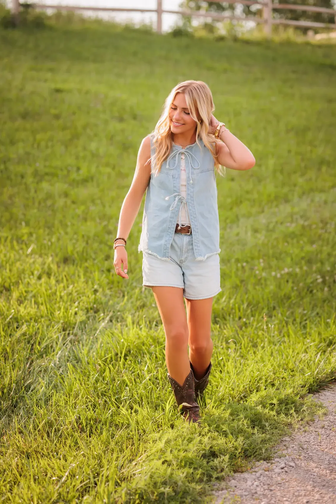 Senior girl with blonde hair wearing a sleeveless denim vest, white top, light denim shorts, and cowboy boots walking on grass on a ranch.