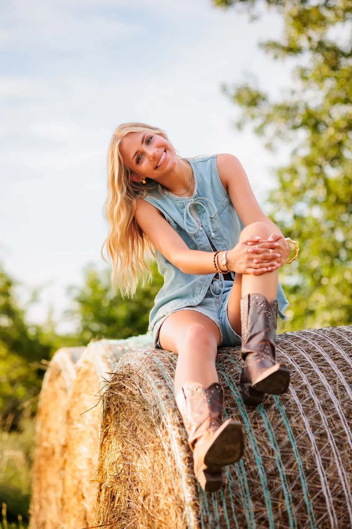 Senior girl with long blond hair wearing a sleeveless denim top, shorts, and cowboy boots, sitting on a large hay bale outdoors with a background of green trees and blue sky.