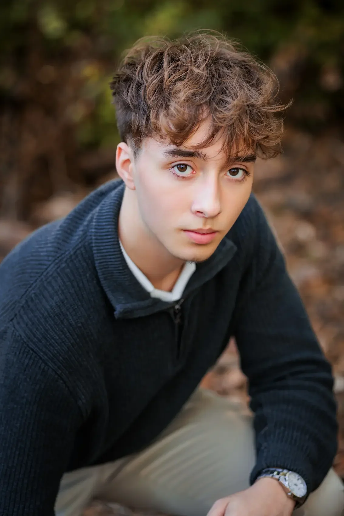 High school senior boy crouching outdoors in Des Moines, looking into the camera, with trees and ground around him.
