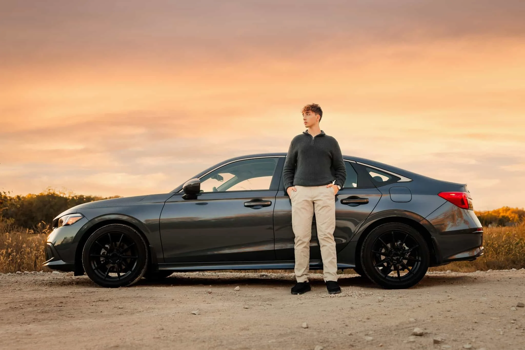 Senior boy standing next to his car on a gravel road at sunset.